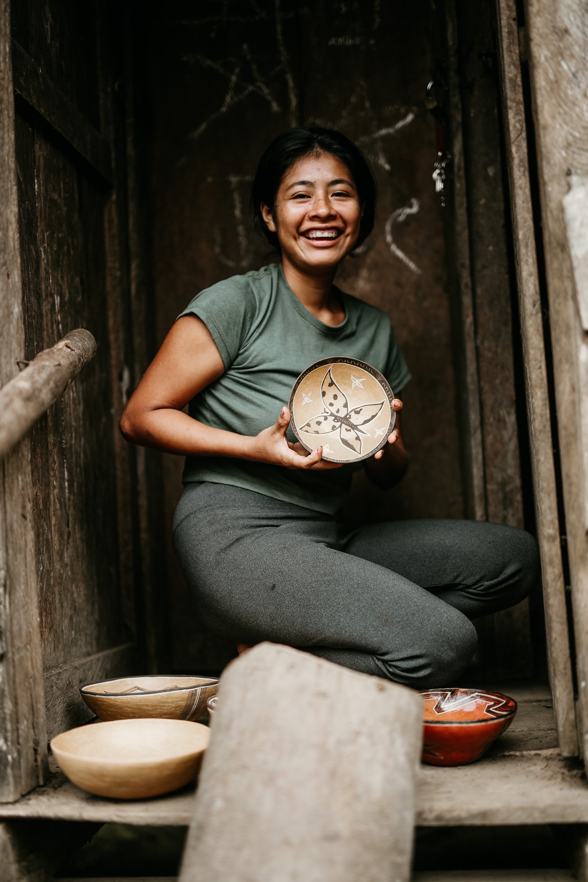 Young woman smiling and kneeling inside a small wooden structure, holding a decorative ceramic plate with a butterfly design, surrounded by other bowls and stones.