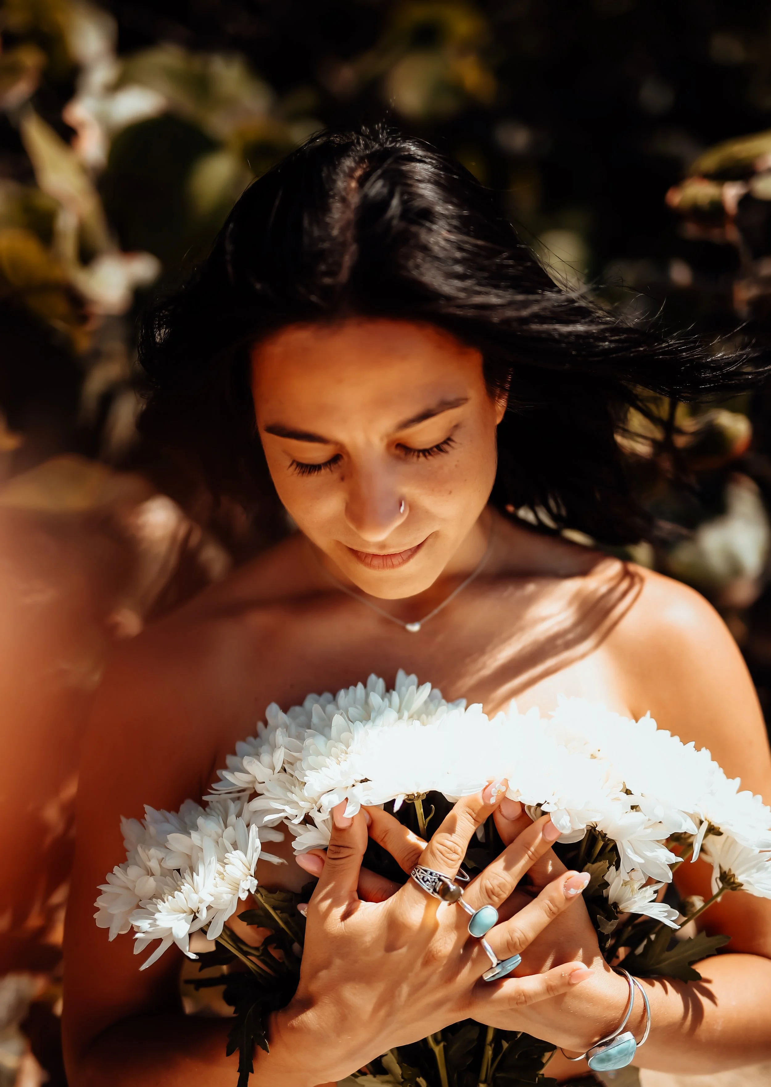 A woman with dark hair holding a bouquet of white daisies, eyes closed, smiling softly, lying on the ground among greenery and flowers.