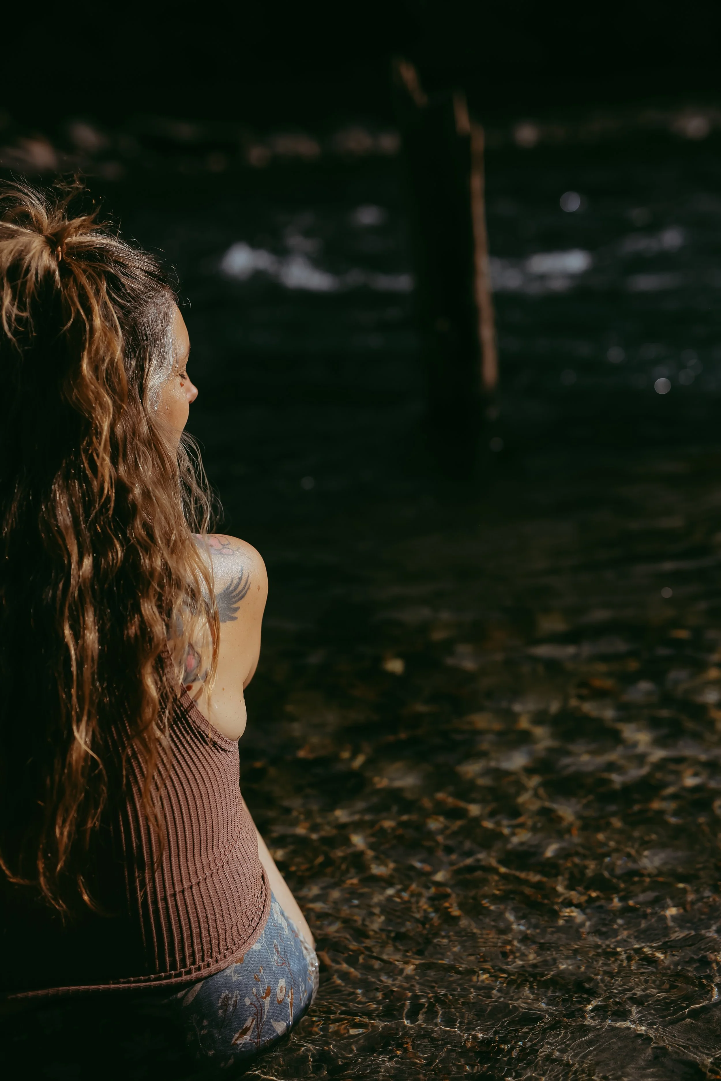 A woman with long hair sitting in shallow water at a beach, facing away from the camera and looking towards the ocean.