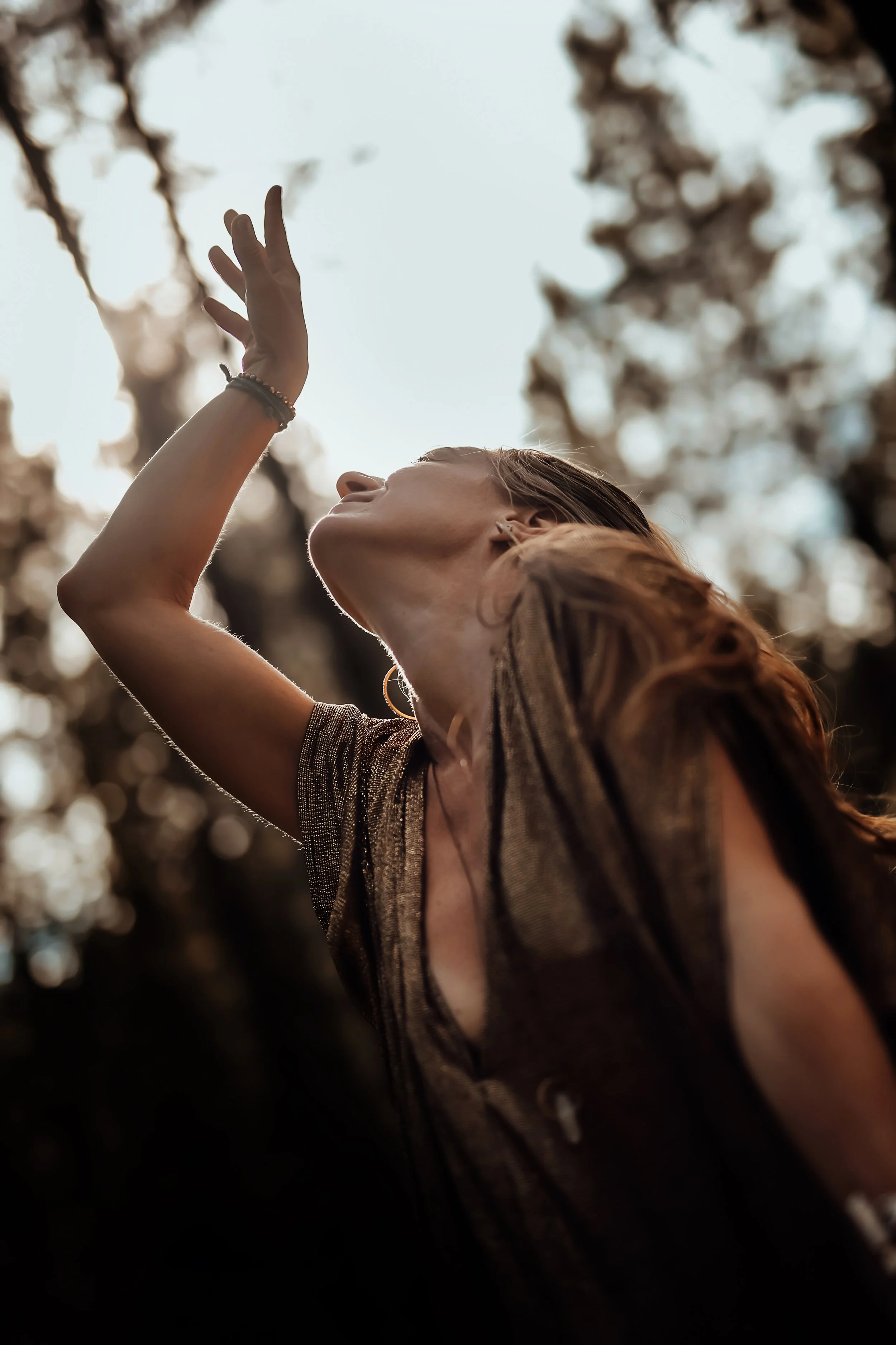 A woman with long hair, wearing a shimmery top and jewelry, is outside with her hand raised, head tilted back, appearing to enjoy the sunlight through blurred trees.