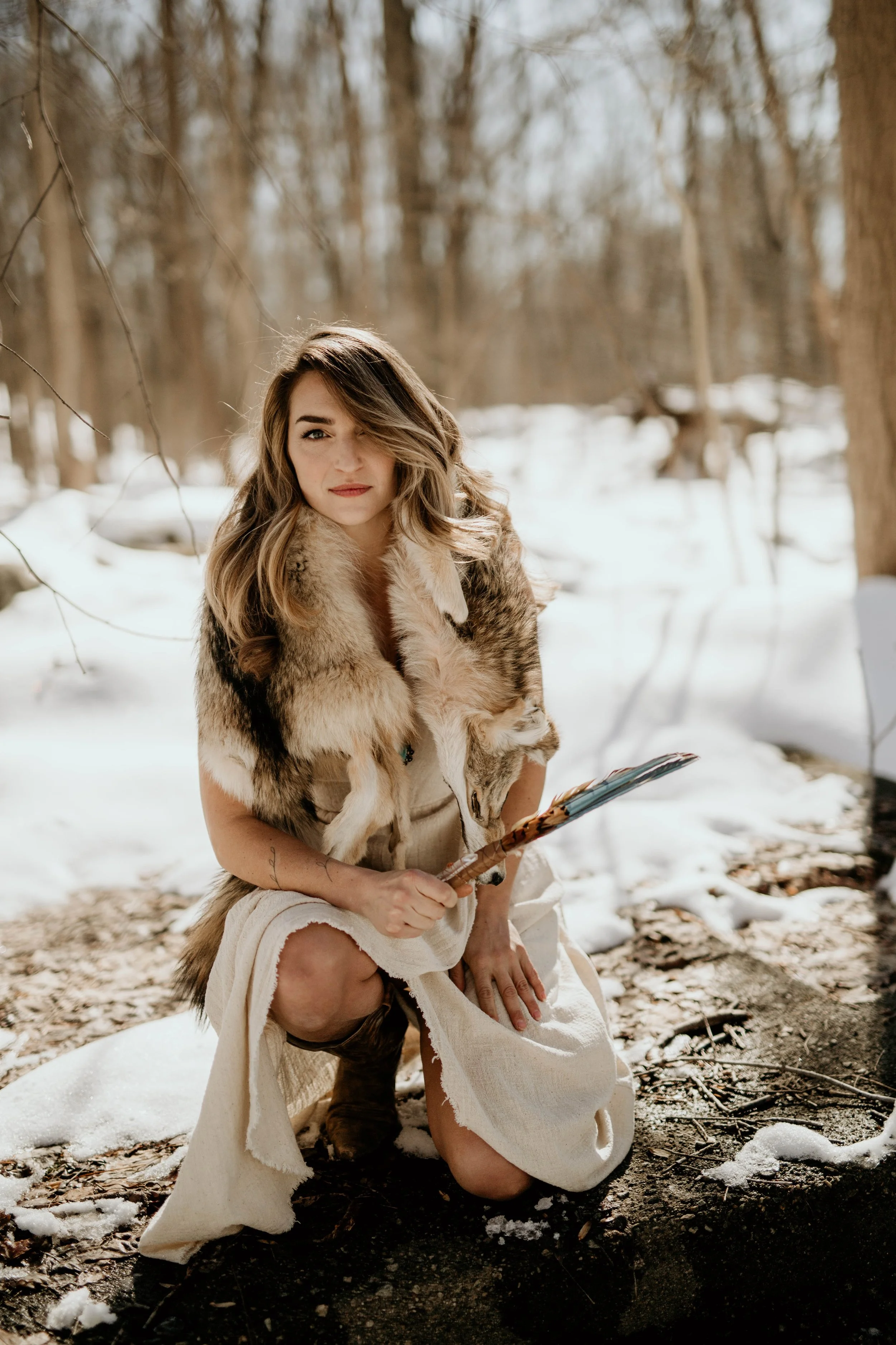 A woman dressed in primitive clothing with a fur cloak kneels outdoors in a snow-covered forest, holding a feathered arrow or spear.