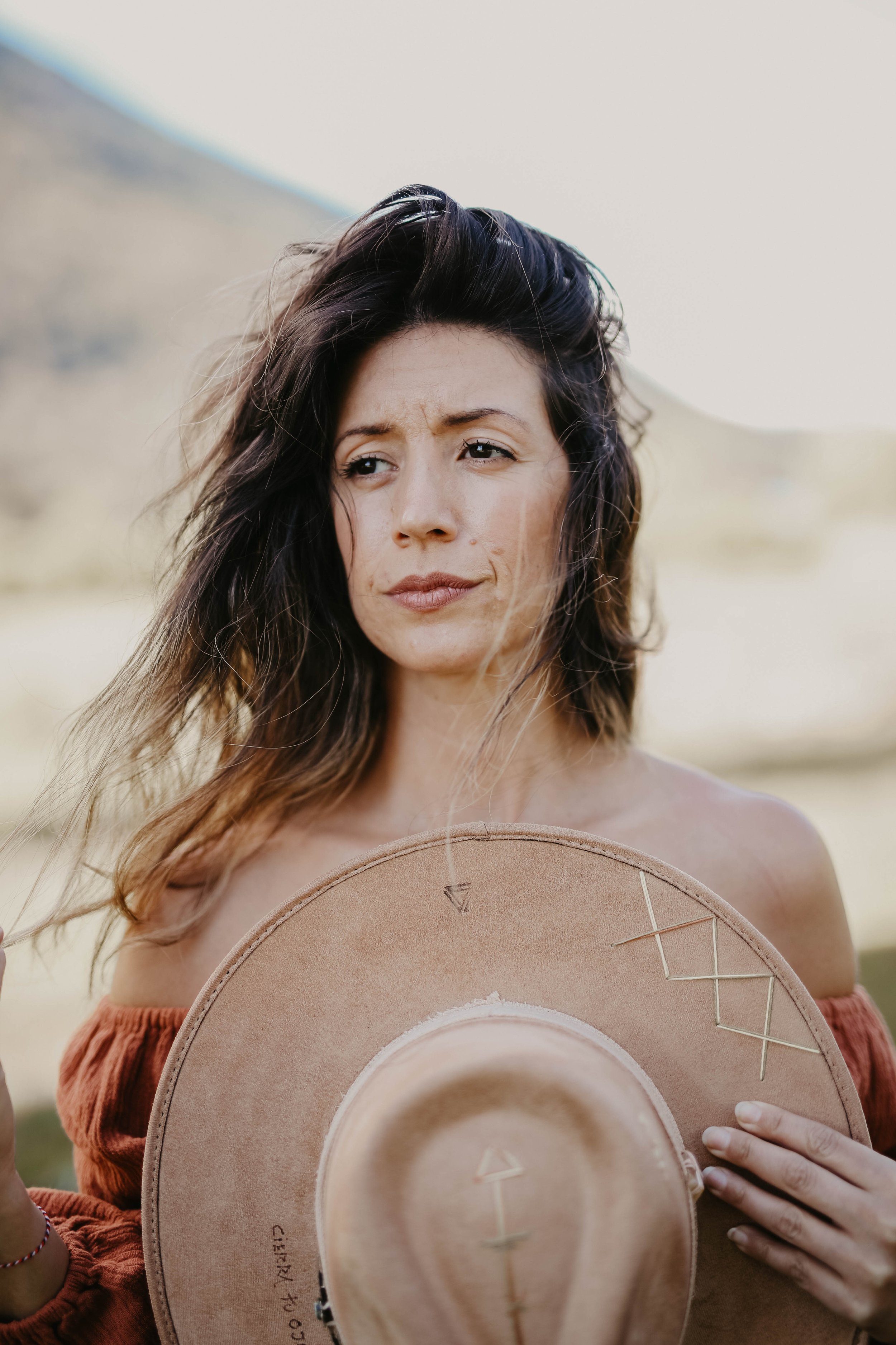 A woman with long, dark, wavy hair holding a wide-brimmed tan hat outdoors. She is wearing an off-the-shoulder, ruffled, reddish-brown top and has a thoughtful or contemplative expression on her face, with a blurred landscape of hills behind her.