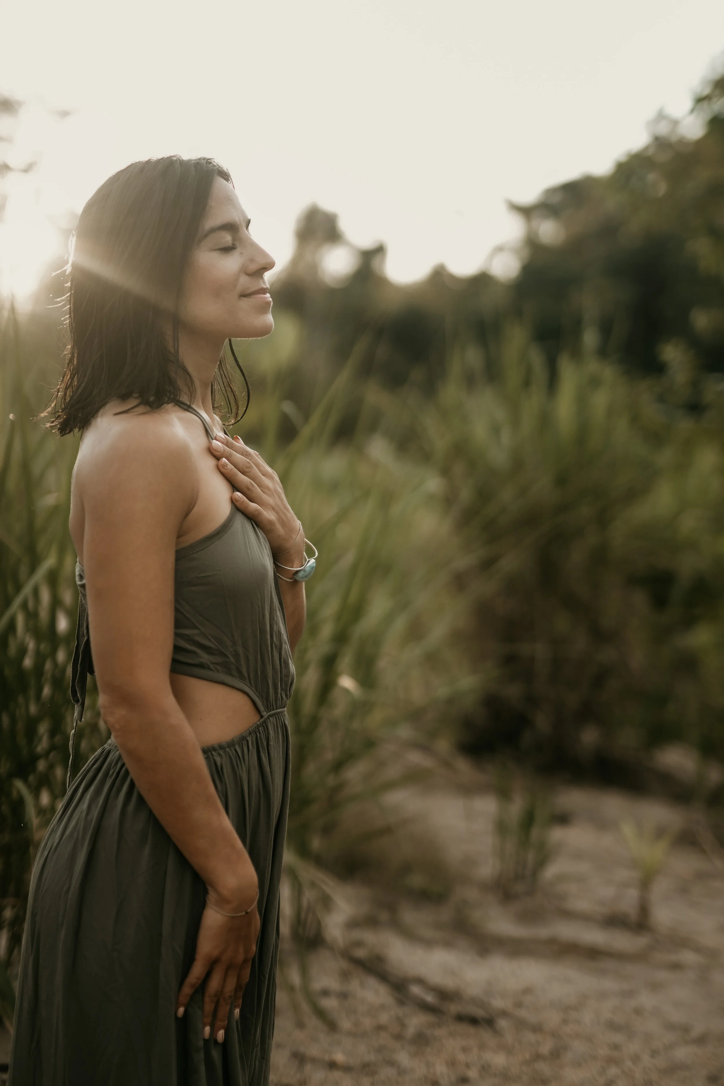 A woman standing outdoors in a field with tall grass, eyes closed, smiling gently, with her hand on her chest, enjoying the moment in the sunlight.