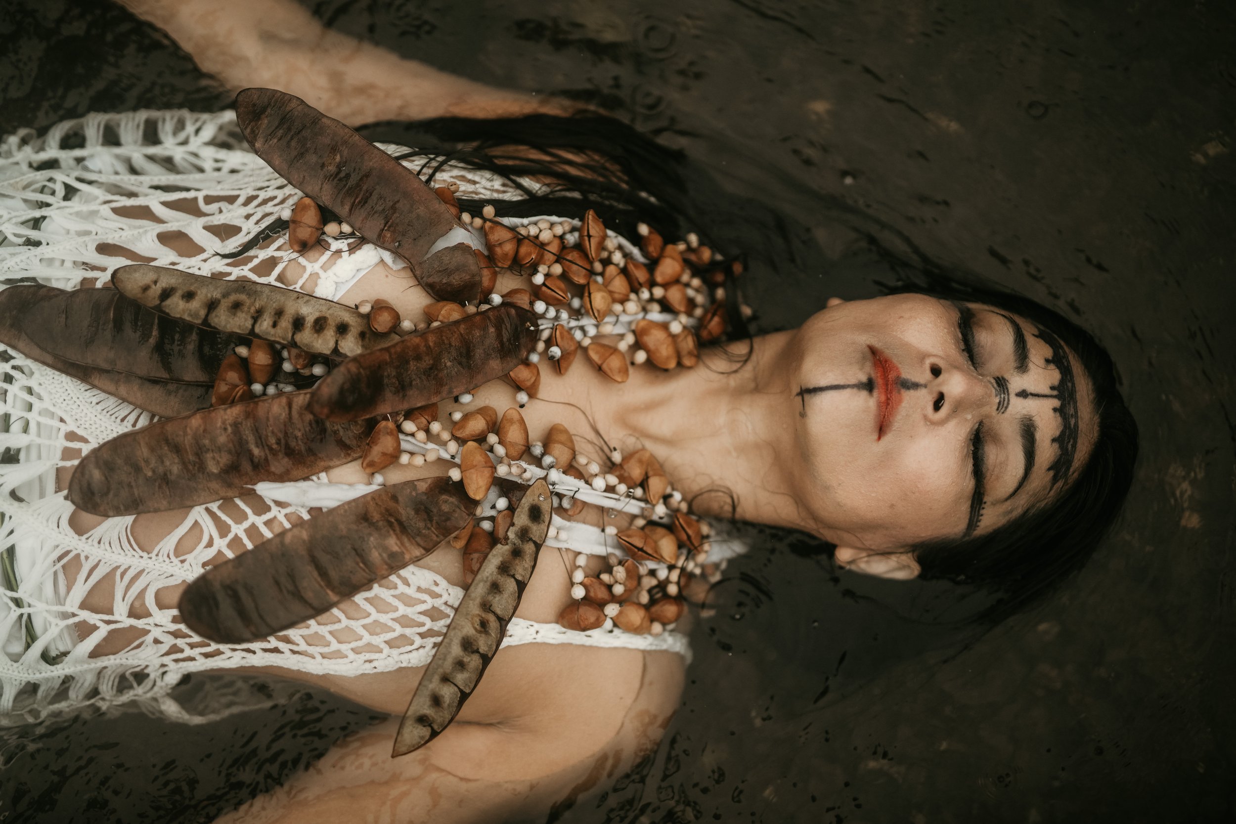 A woman lying in water with her eyes closed, wearing a crochet top, adorned with large dark feathers and beads on her chest, and makeup resembling a tribal or ceremonial look.