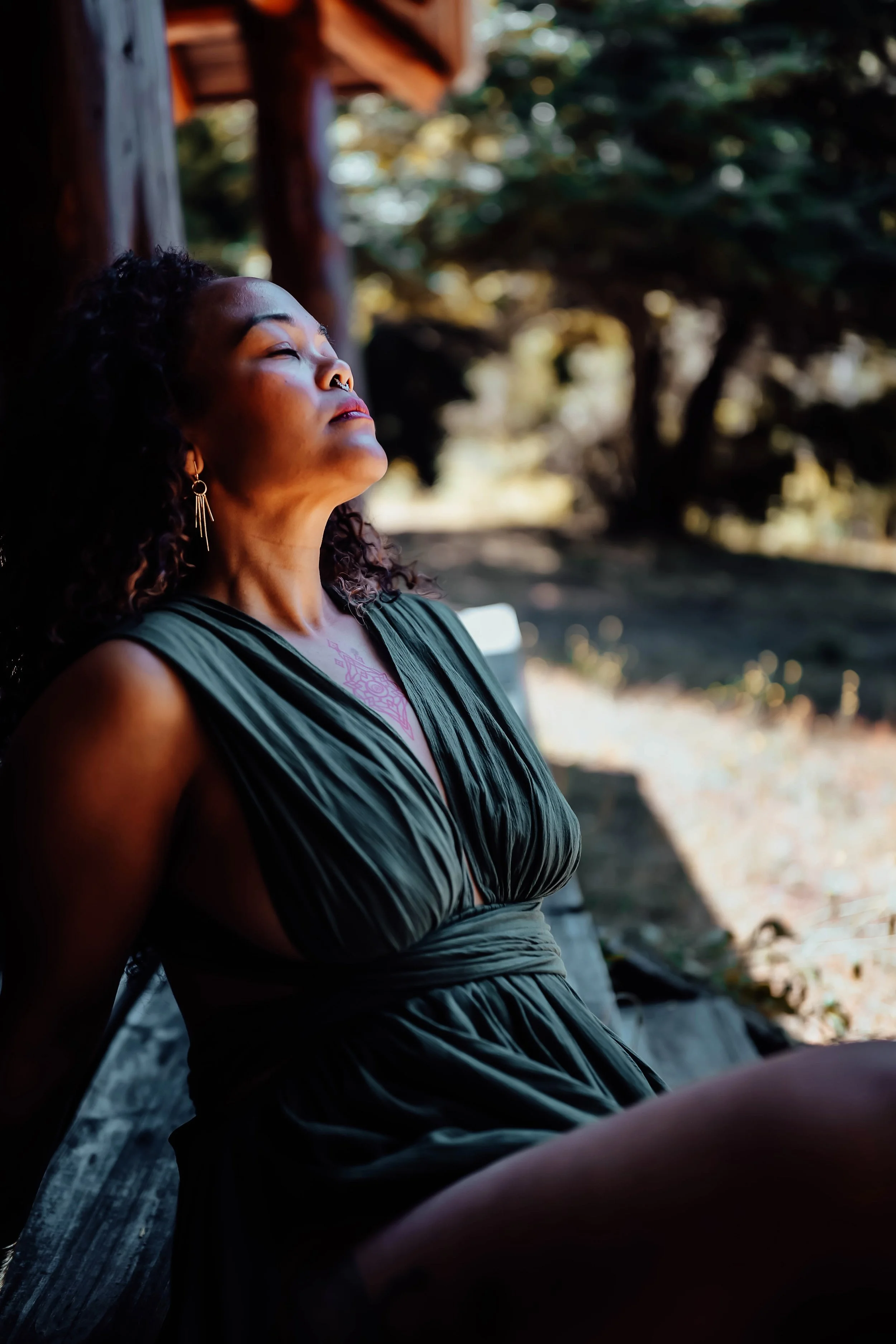 A woman with curly hair and a nose ring is sitting outdoors with her eyes closed, wearing a dark green sleeveless dress, and leaning against a wooden structure in a natural setting.