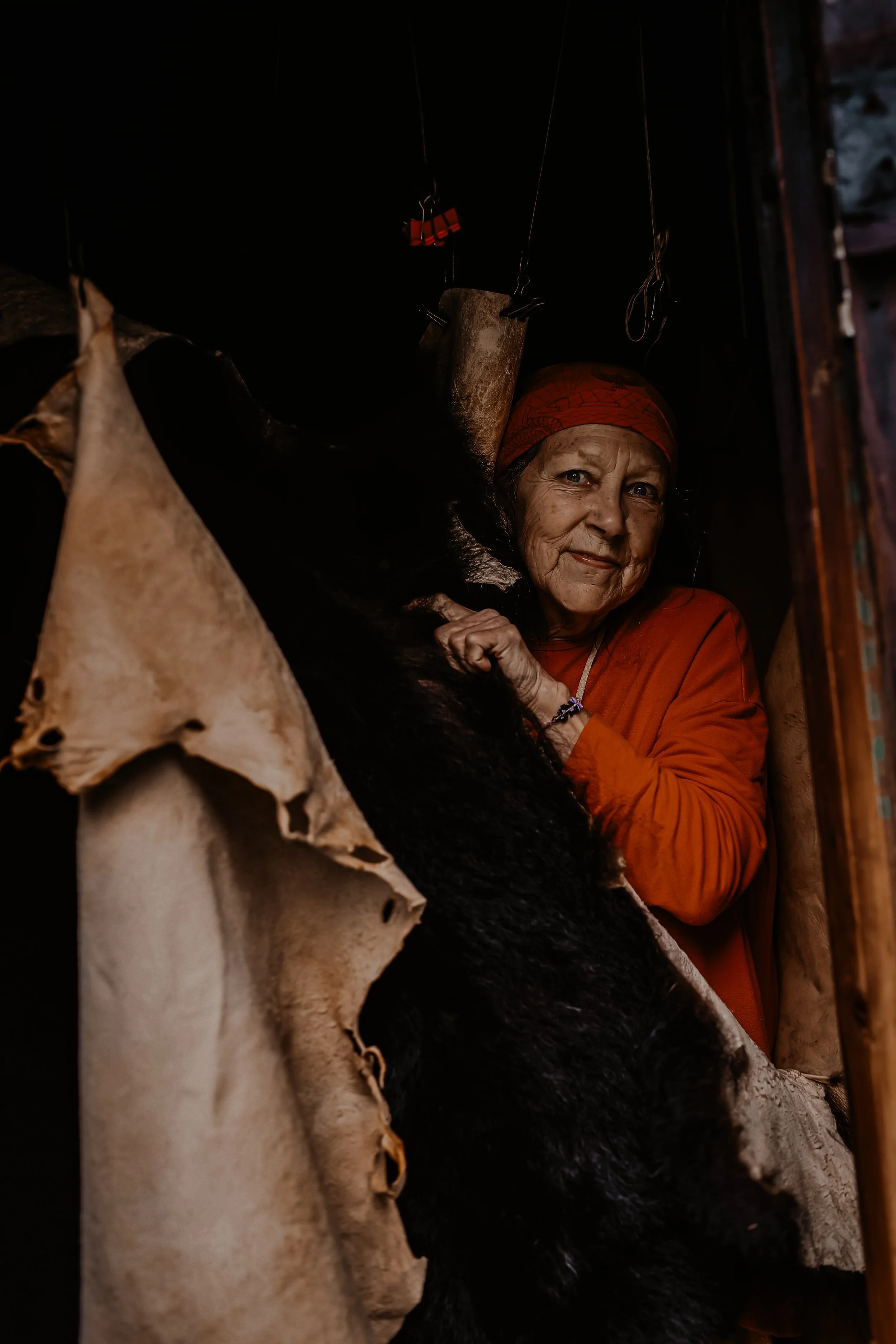 An elderly woman in an orange headscarf and shirt inside a dark room, looking at the camera, with a black bear and animal skins hanging around her.