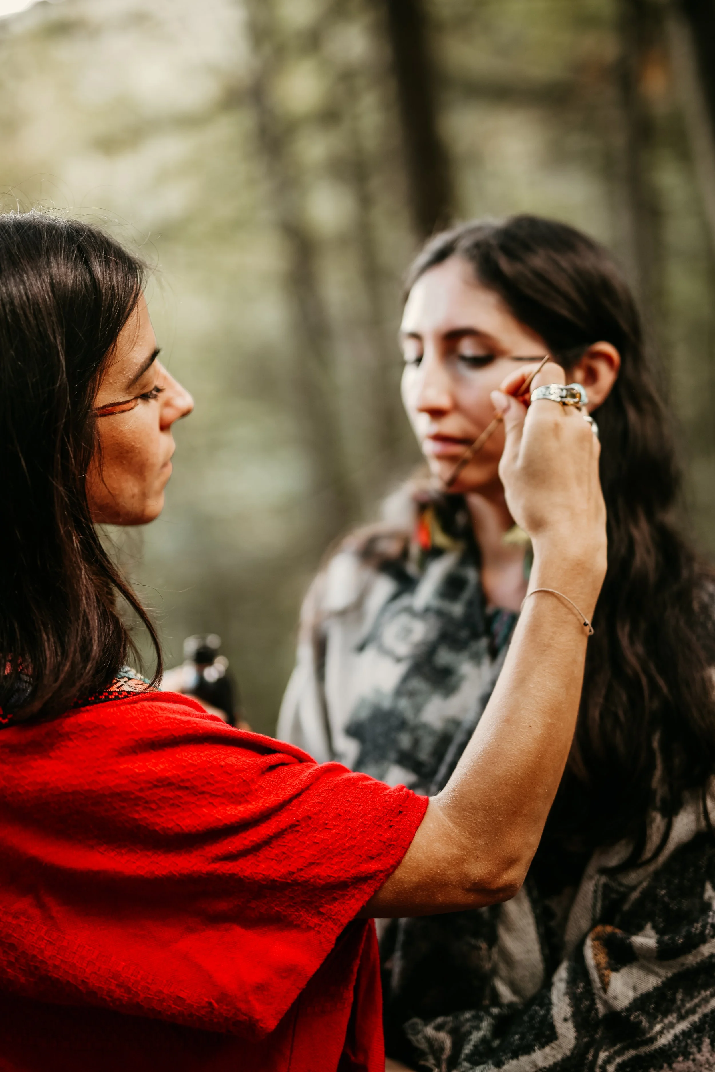 A woman in a red shirt applying makeup to another woman with dark hair in an outdoor wooded setting.