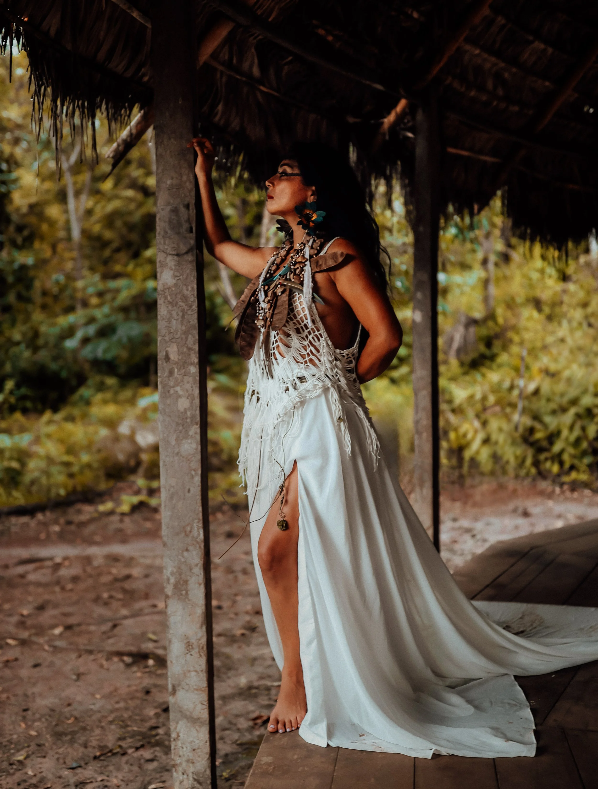 Woman in bohemian style dress standing on a wooden platform under a thatched roof, with a tropical background of trees and foliage.