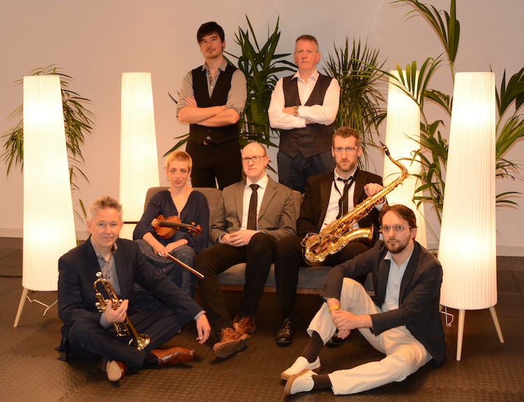 Seven musicians, in a room with large potted plants and tall, white floor lamps.
