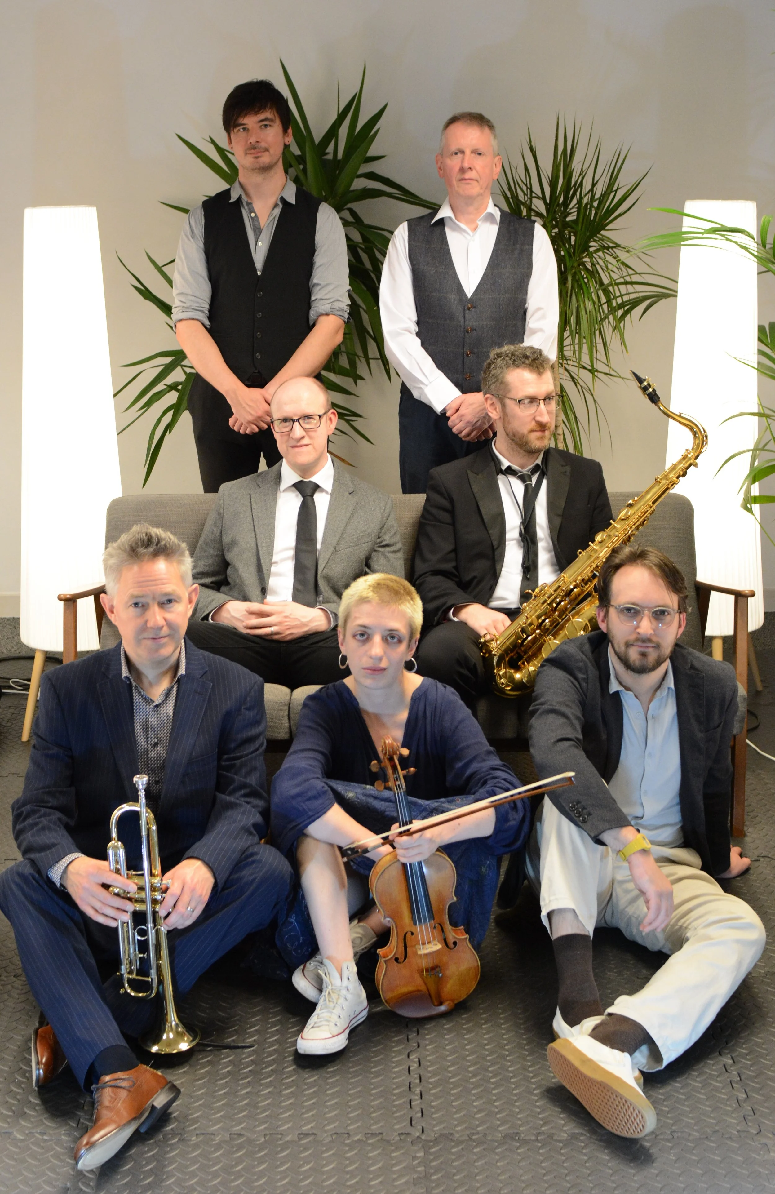 Seven musicians posing for a photo indoors, with three holding instruments: tenor saxophone, violin and trumpet. In the background are large green plants and modern white lamps.