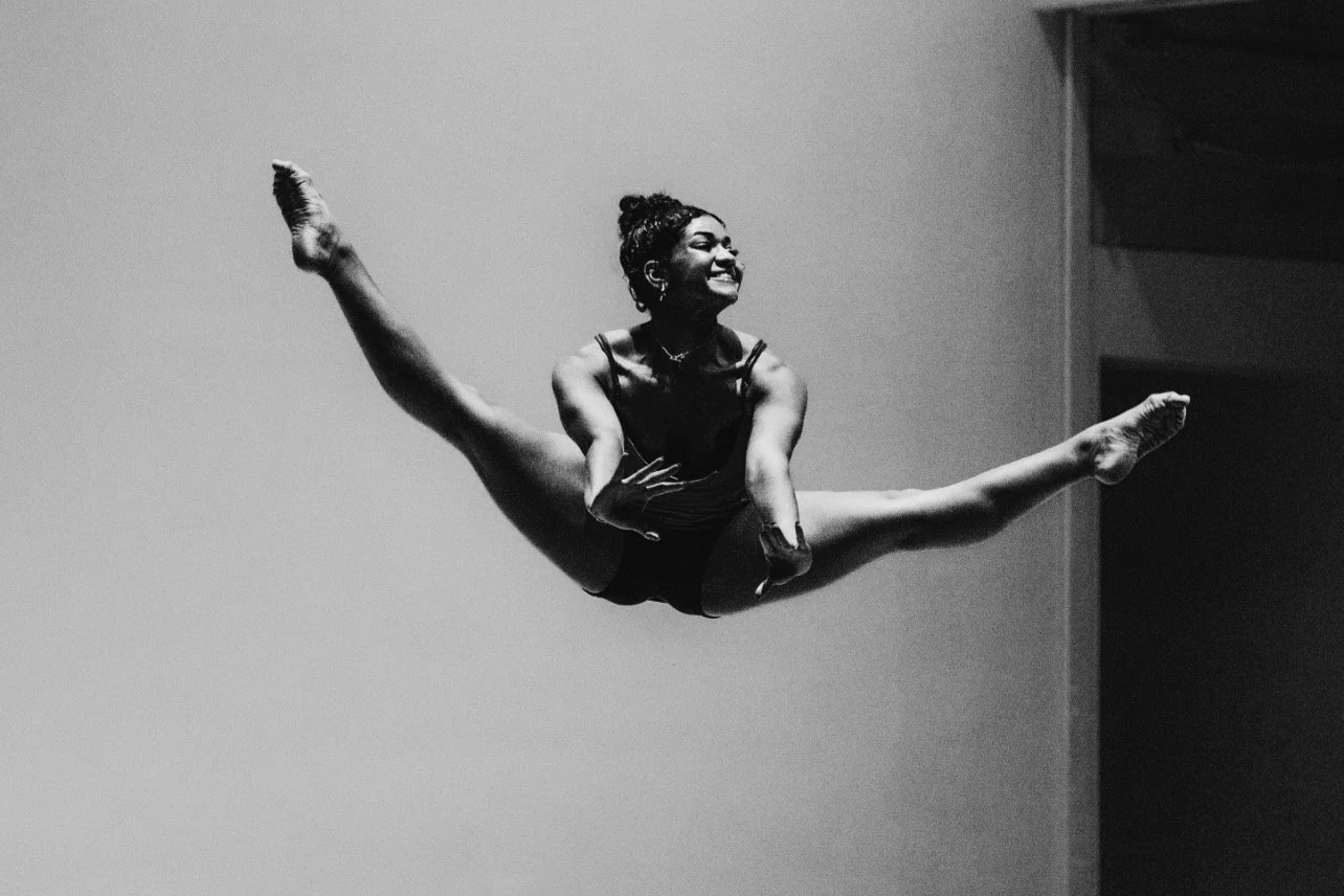 Black and white photo of a young woman performing a split leap in a gym or dance studio with a plain wall background, smiling and looking upwards.