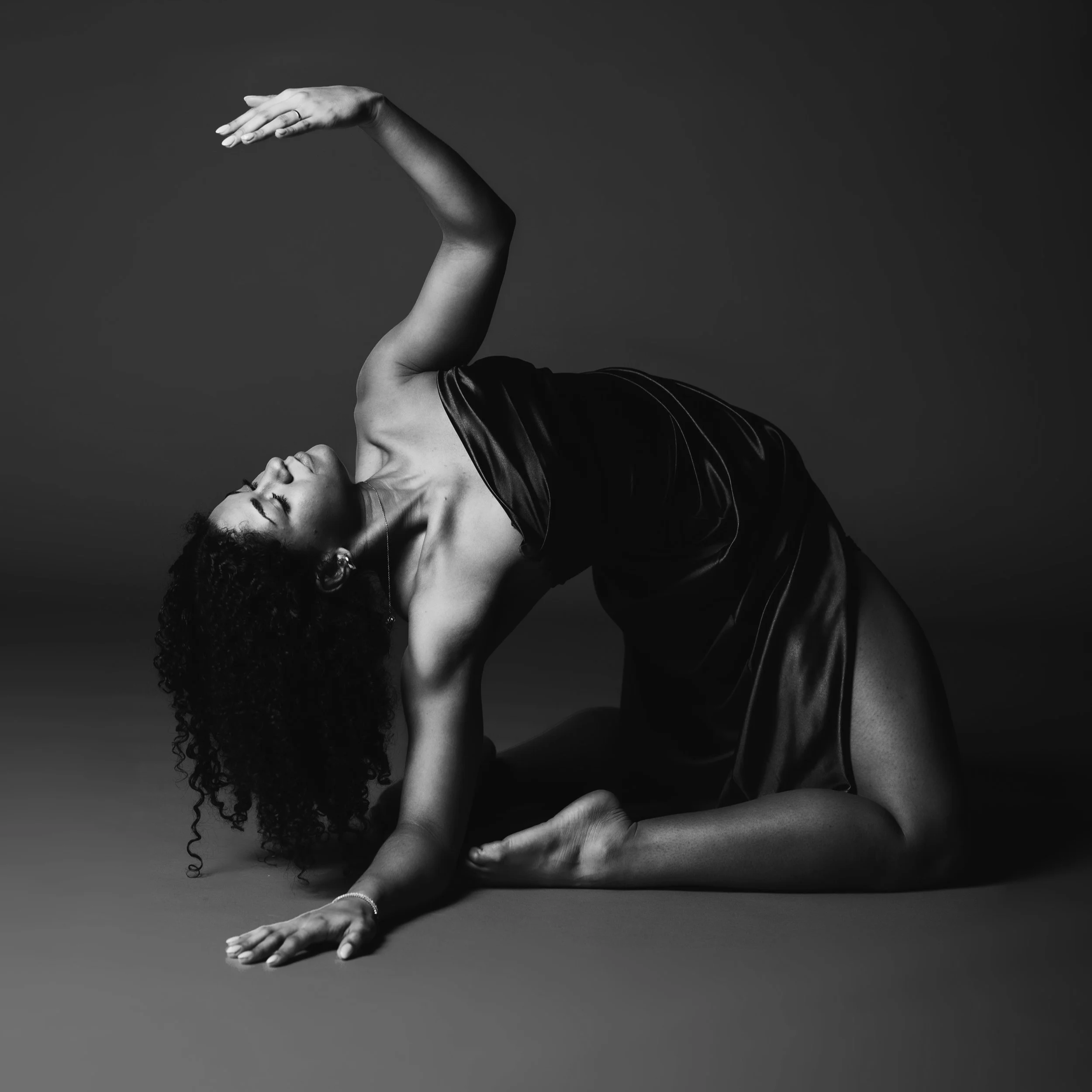 Black and white photo of a woman doing a yoga pose on the floor, wearing a shiny dress, with her head tilted back and one arm stretched overhead.