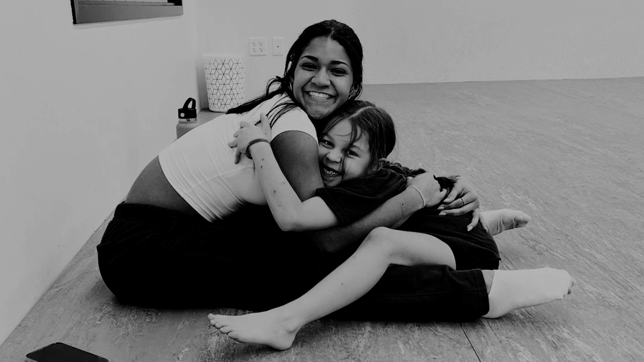 A woman and a young girl sitting on the floor hugging and smiling at the camera, in a room with minimal decor.