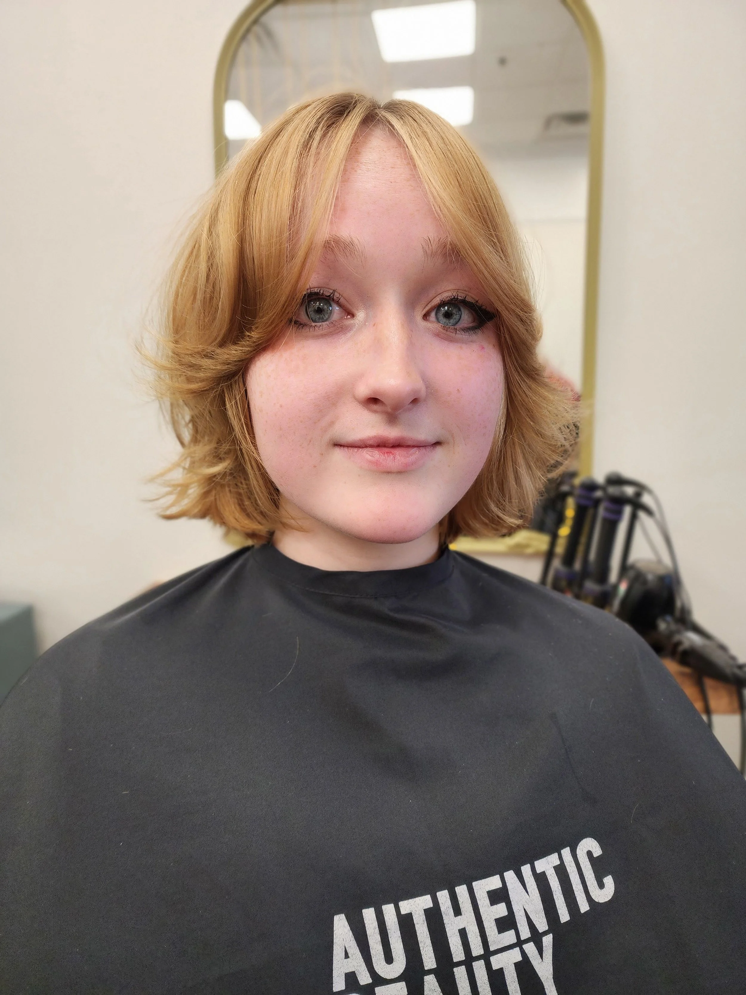 A young woman with shoulder-length red hair, blue eyes, and freckles, looking at the camera in a salon or studio, wearing a black cape with the word 'AUTHENTIC' printed on it, with salon tools and a mirror in the background.