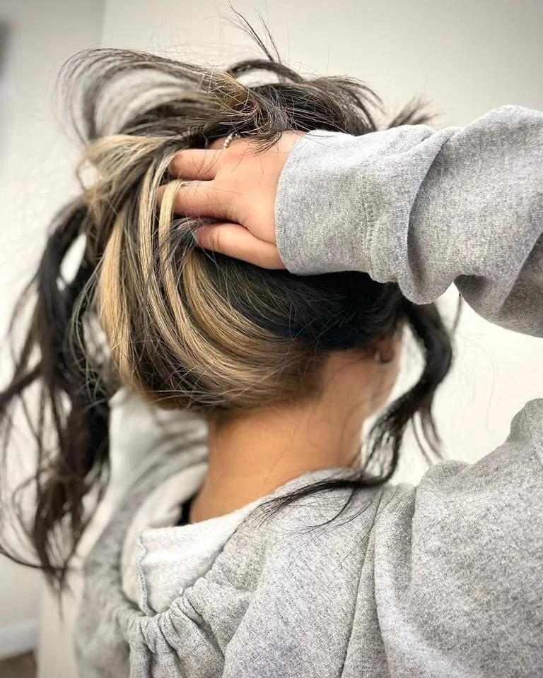 Woman with long dark hair with blonde highlights, wearing a gray hoodie, holding her head with both hands.
