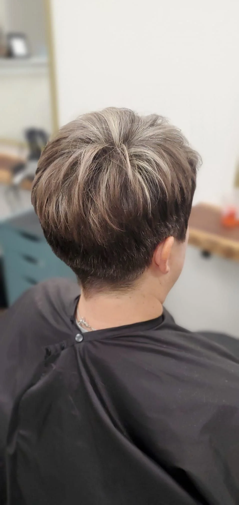 Back view of a person with a stylish short layered haircut with highlights, sitting in a salon chair.