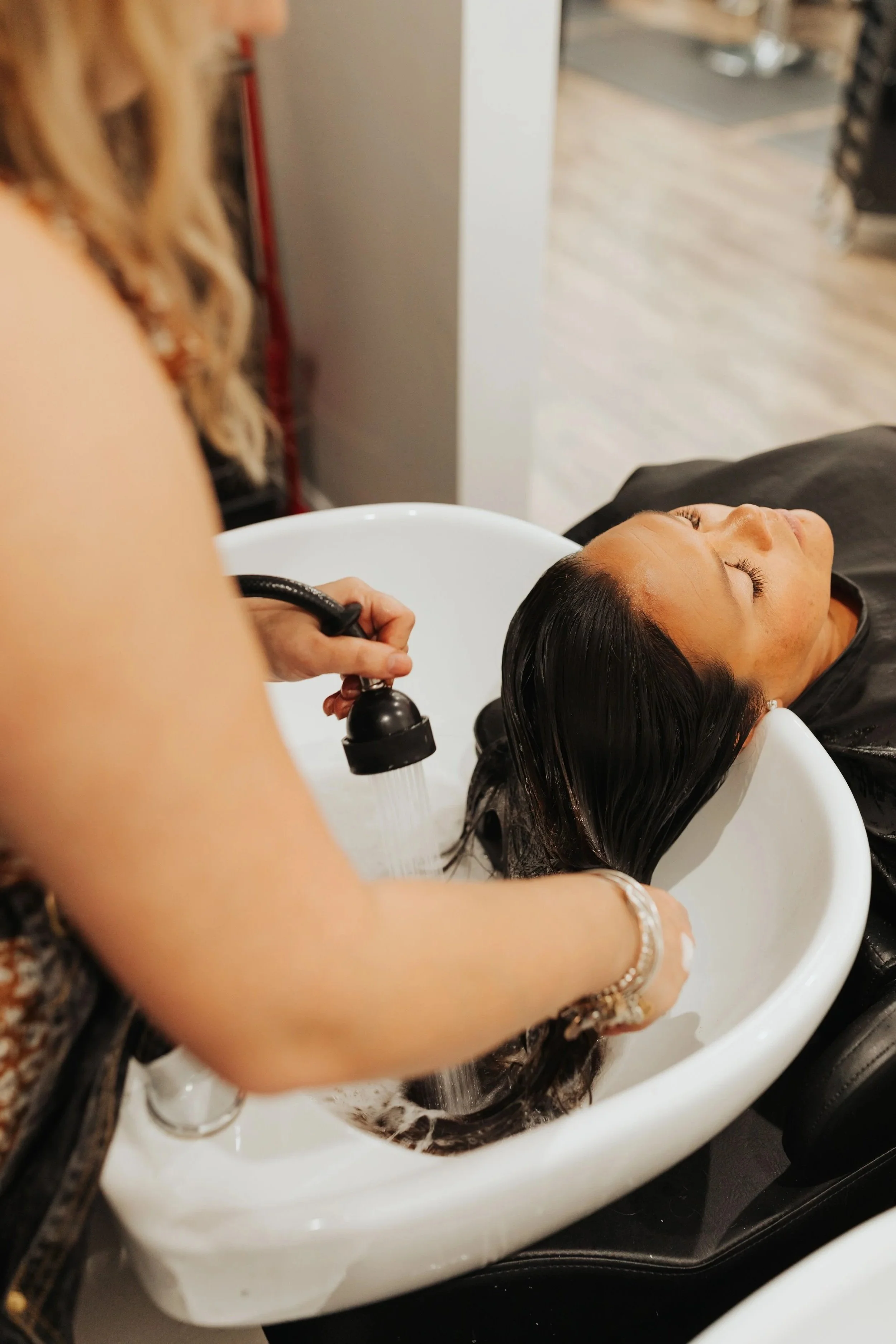 A woman is having her hair washed at a salon while lying back in a shampoo bowl, with another person washing her hair using a spray nozzle.