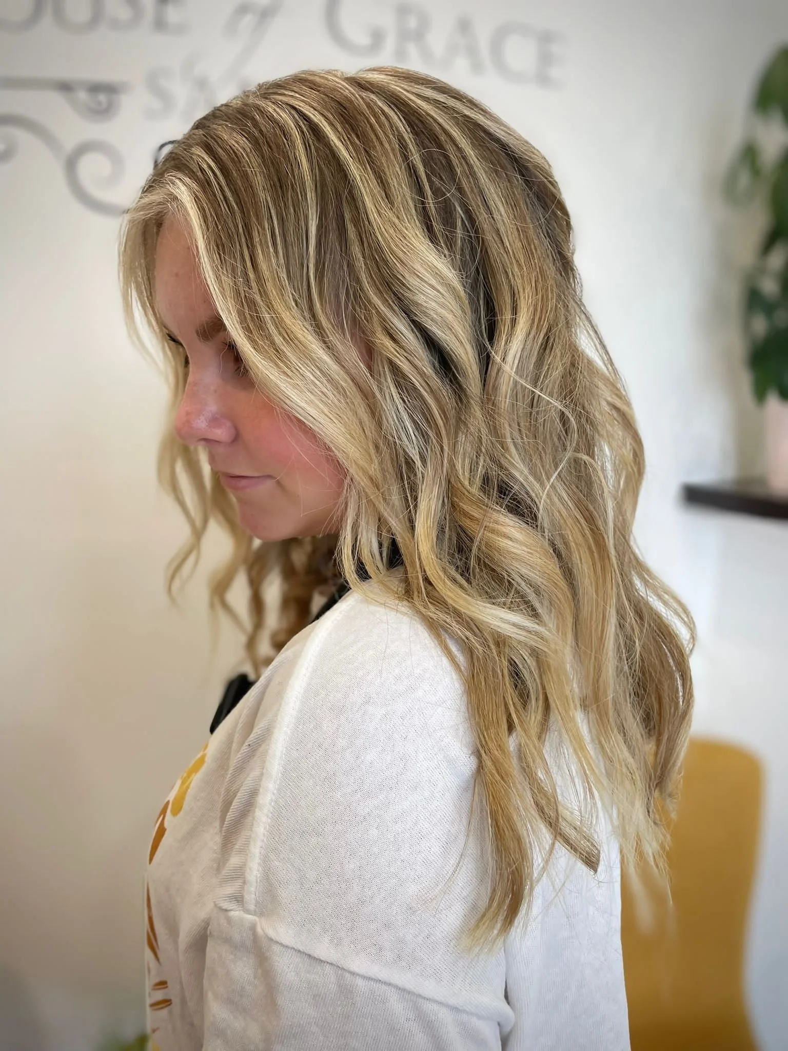 Side view of a woman with long, wavy blonde hair, wearing a light-colored sweater, standing indoors with a wall and a plant in the background.