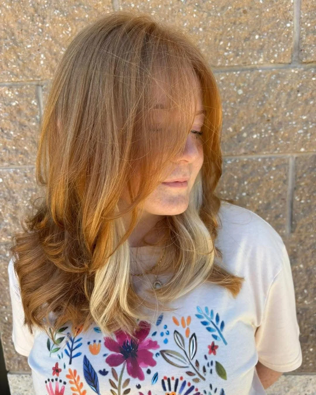 A woman with long, wavy, light brown hair with blonde highlights, looking down, standing against a tan brick wall. She is wearing a white T-shirt with colorful floral prints.