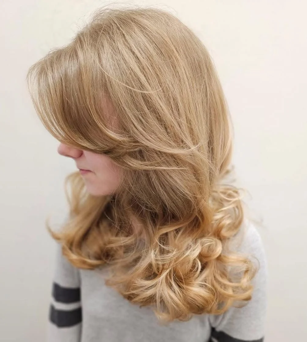 Side view of a woman with shoulder-length, blonde, curly hair wearing a gray top with black and white striped sleeves.