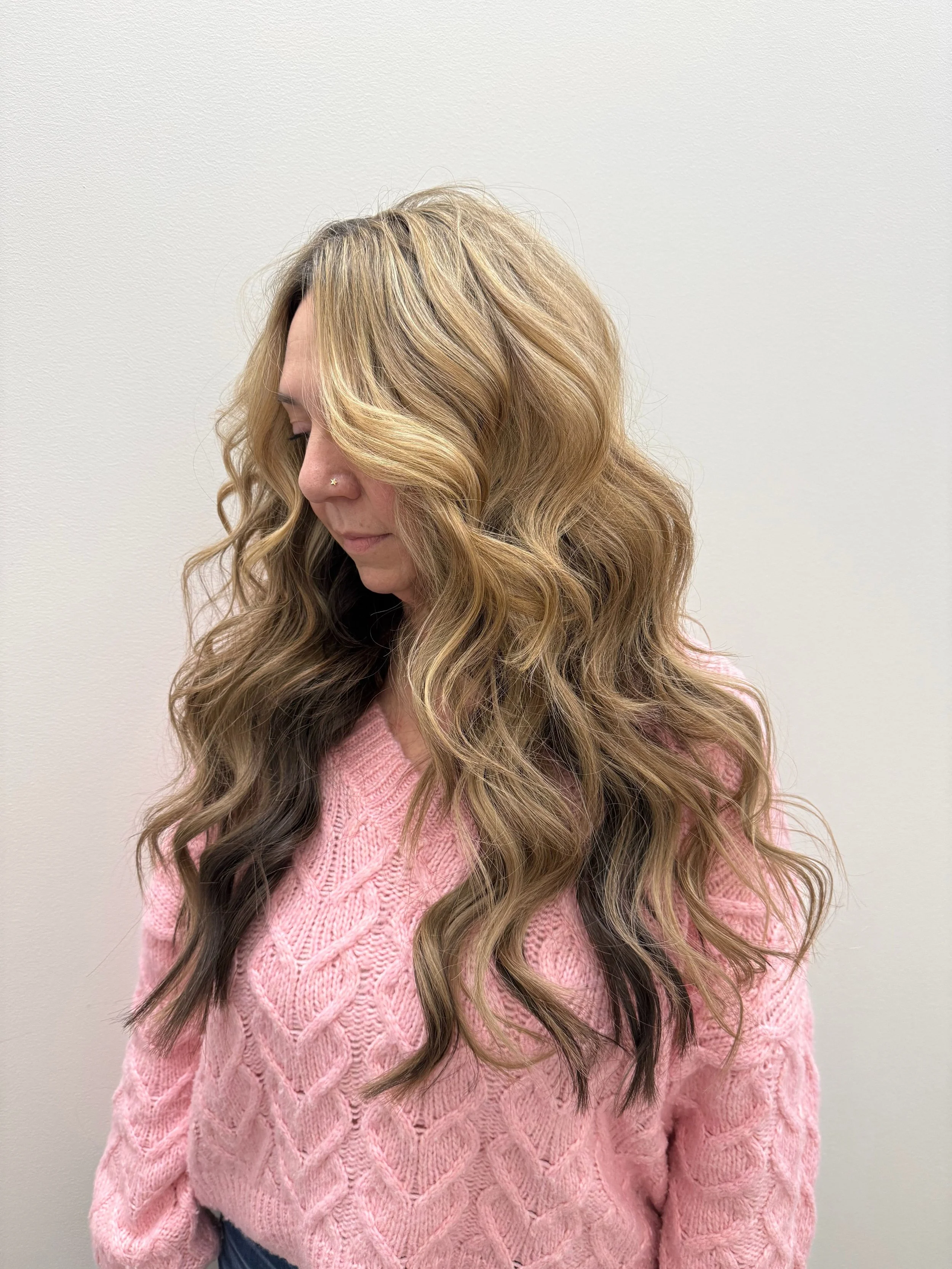 A woman with long wavy hair dyed with blonde highlights and darker lowlights, wearing a pink cable-knit sweater and standing against a plain off-white wall.