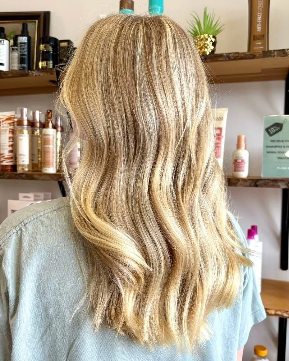 Blonde hair styled in loose waves, seen from the back, in a salon setting with shelves of hair products in the background.