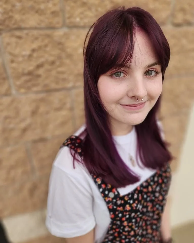 Young woman with purple hair, light skin, a septum piercing, wearing a white t-shirt and a floral dress, standing against a brick wall.