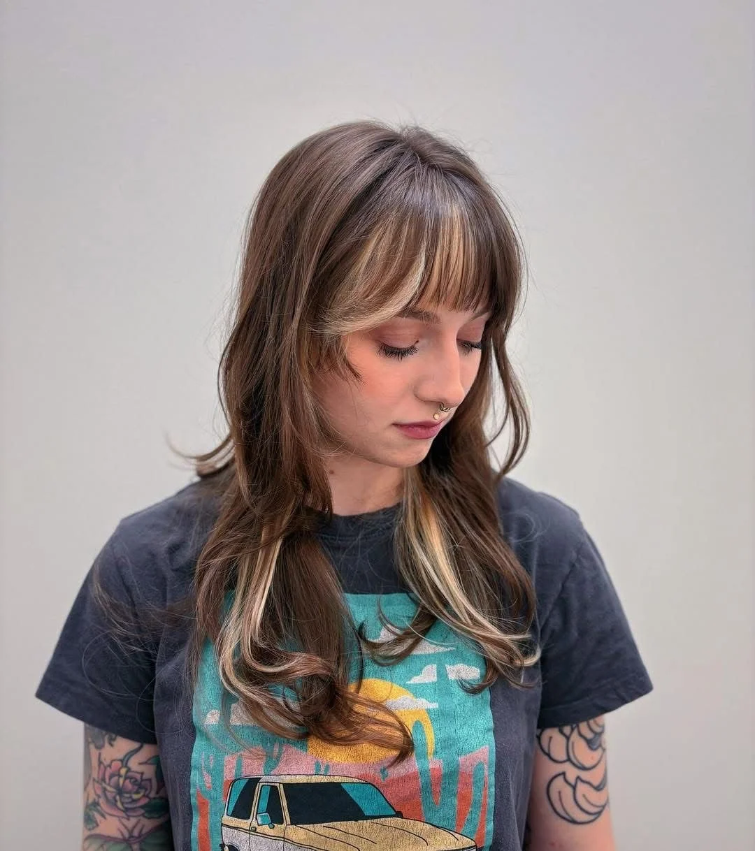 Young woman with long, wavy hair and tattoos on her arms, looking down, wearing a graphic t-shirt with a car and sunset design, in front of a plain gray background.