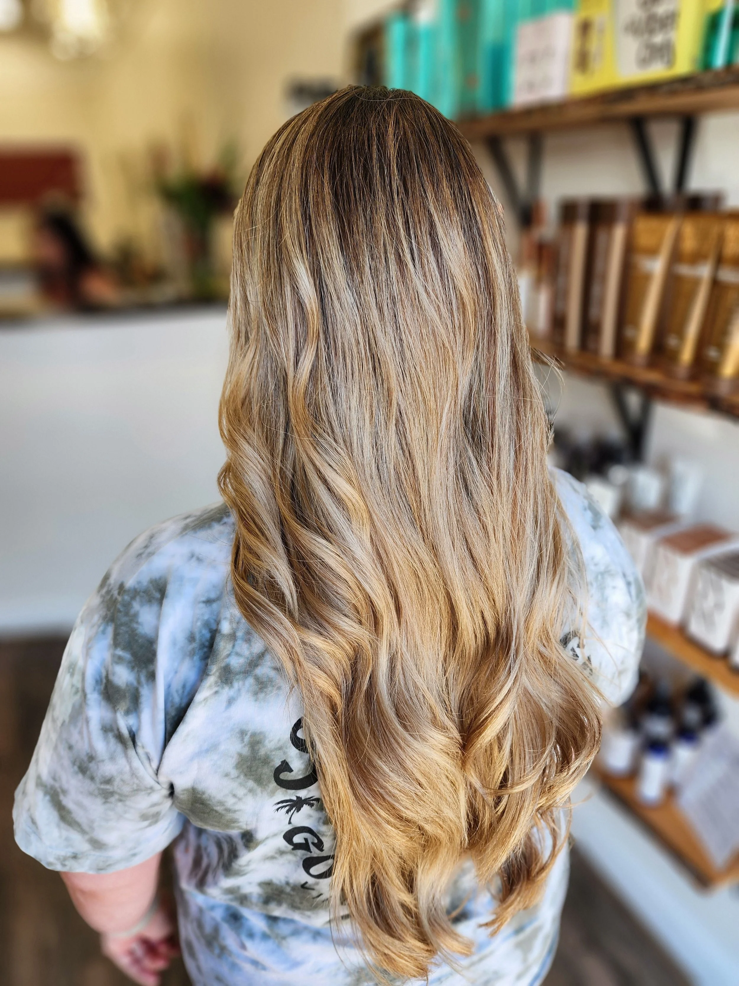 Back view of a person with long, wavy, highlighted blonde hair in a salon or store setting.