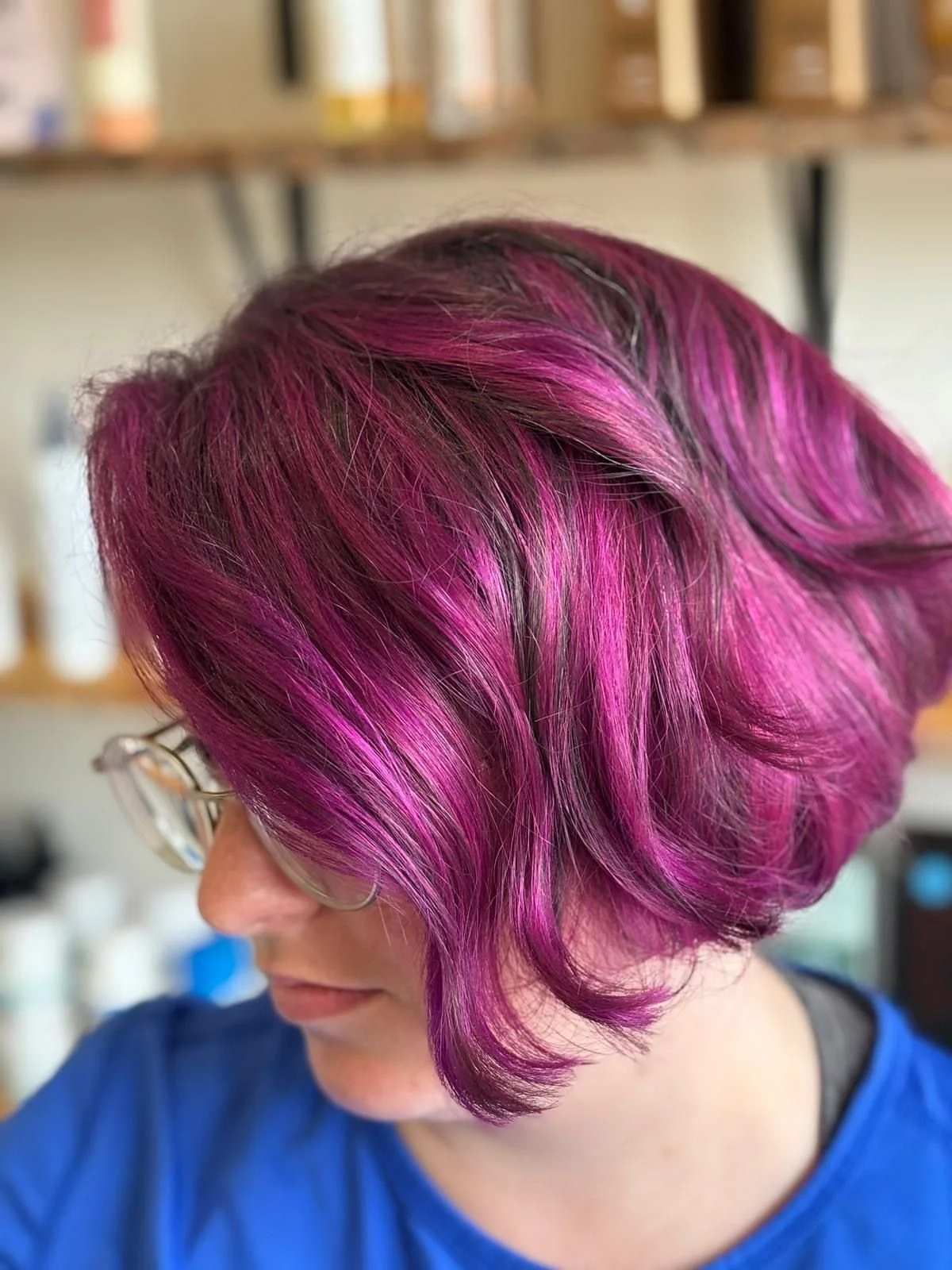 Woman with short, wavy hair dyed in shades of pink and purple, wearing glasses, in a salon