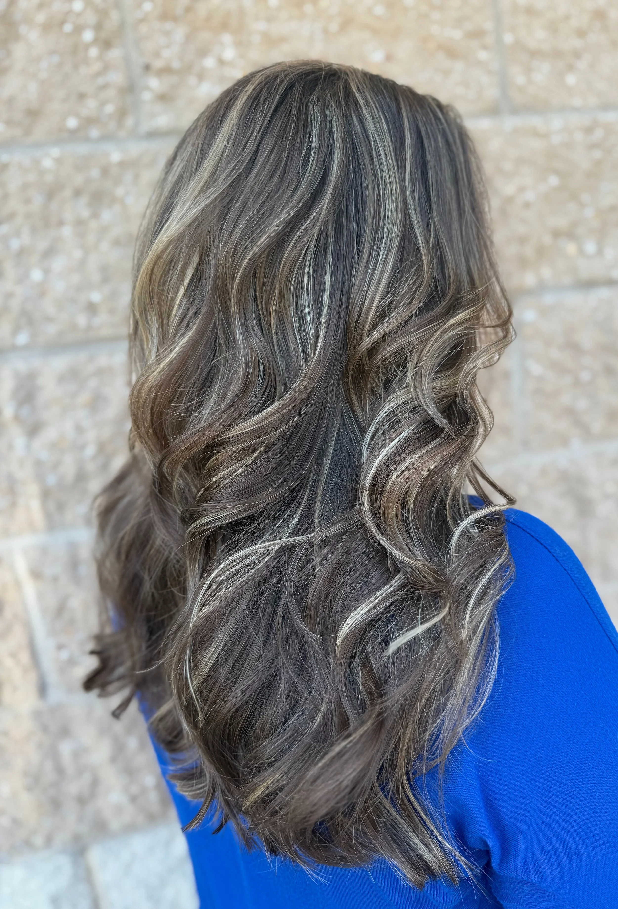 Side view of a woman with long, wavy, gray and blonde highlighted hair, wearing a blue top, with a brick wall background.