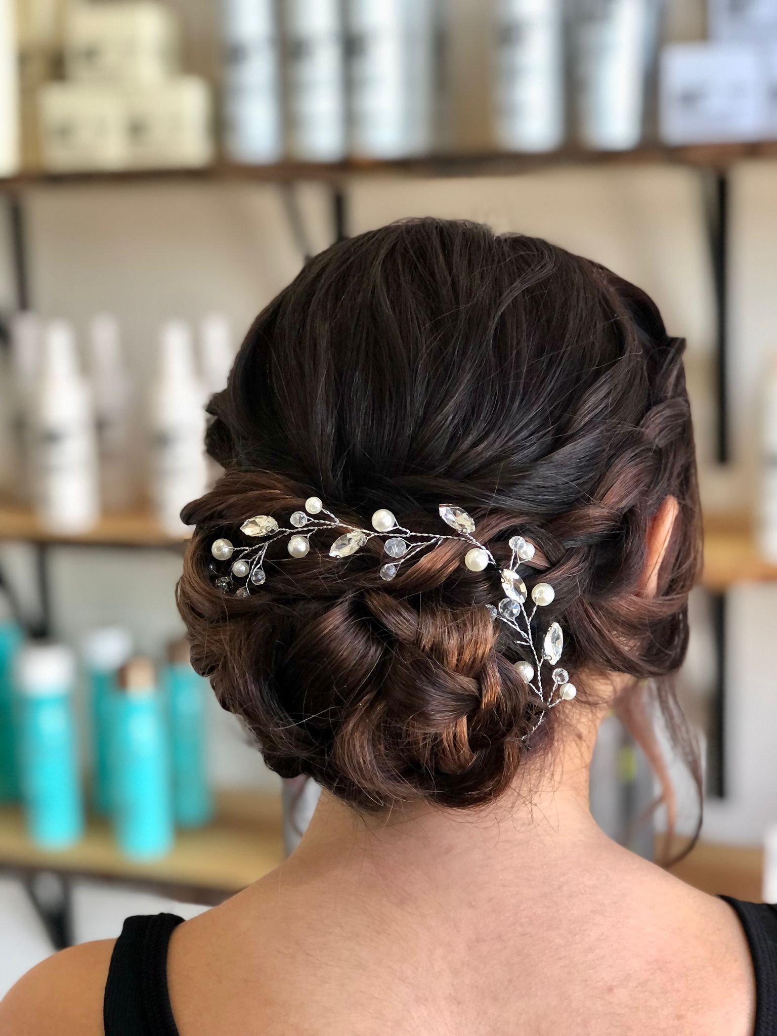 Back of a woman's head with styled dark brown hair in an updo decorated with pearl and crystal hair accessories, set in a salon or studio with shelves of hair products in the background.