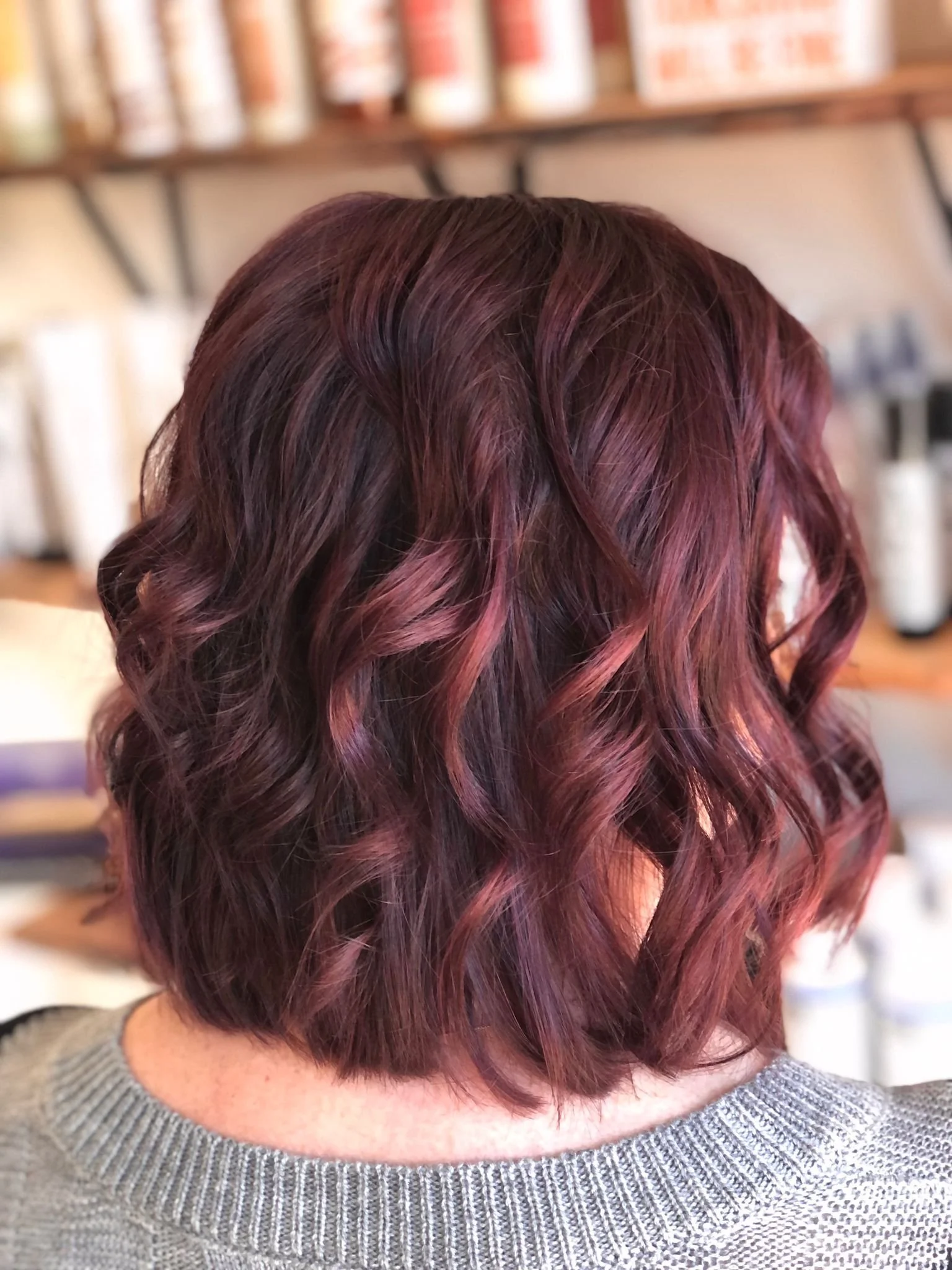 Close-up of a woman's wavy, shoulder-length hair dyed in a blend of deep burgundy and purple shades, with a blurred background of shelves with hair products.
