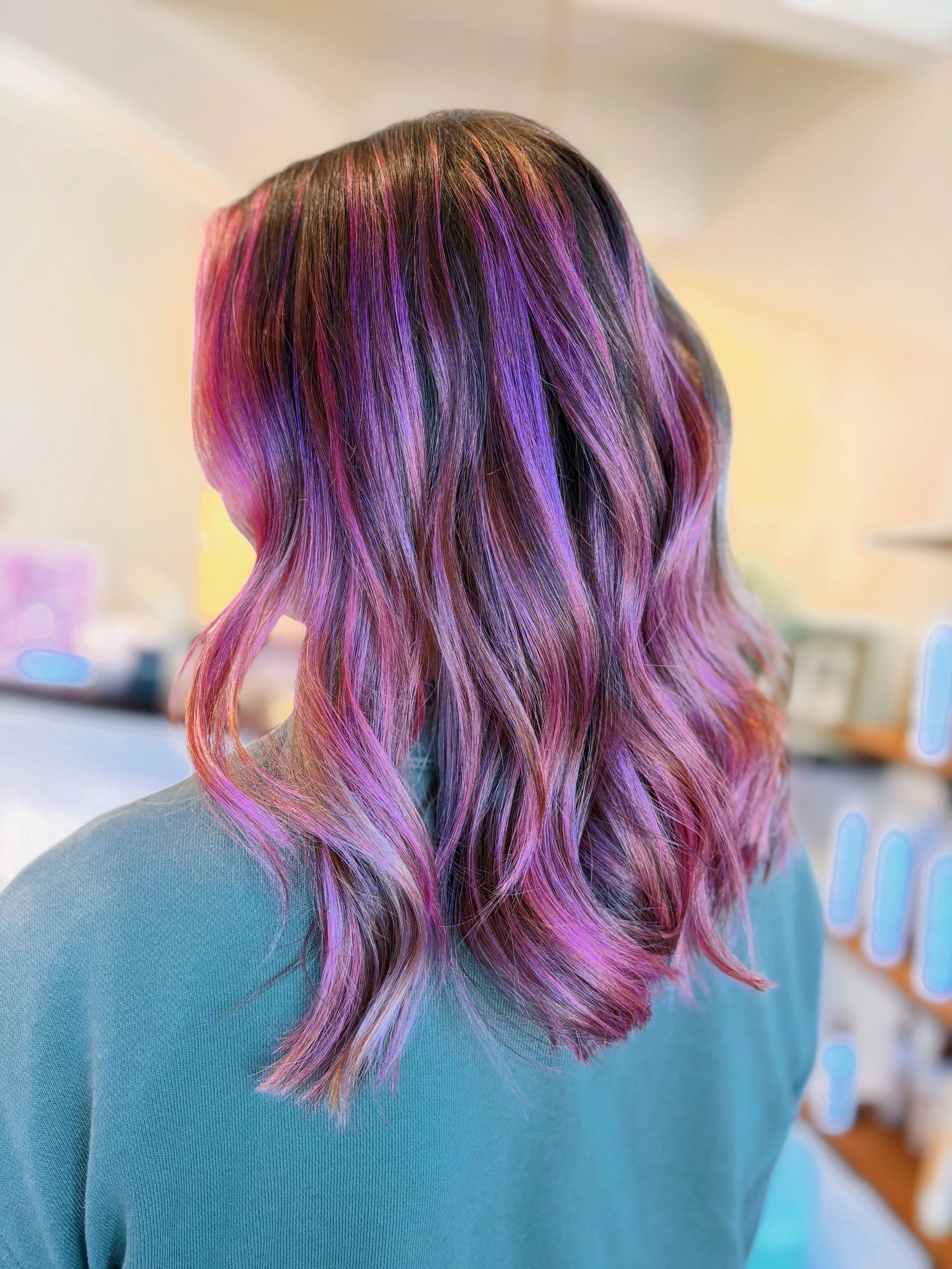 Back of a person's head with wavy hair dyed in shades of purple, pink, and blue, wearing a light blue sweater.