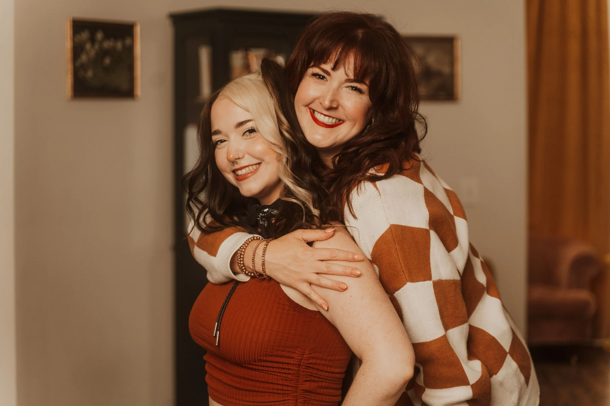 Two women hugging and smiling, one with dark hair and the other with blonde hair, both wearing warm clothing, indoors with framed pictures on the wall in the background.