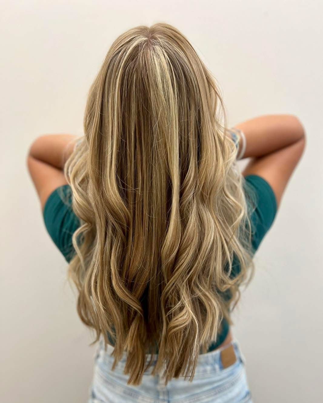 Back view of a woman with long, wavy blonde hair, wearing a teal shirt and light-colored pants, against a plain white wall.