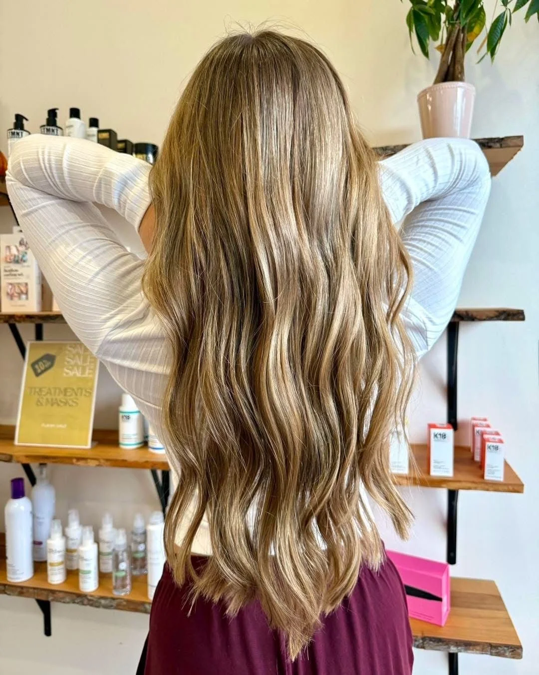 A woman with long, wavy, light brown hair seen from the back inside a salon, with shelves of hair products and a plant in the background.