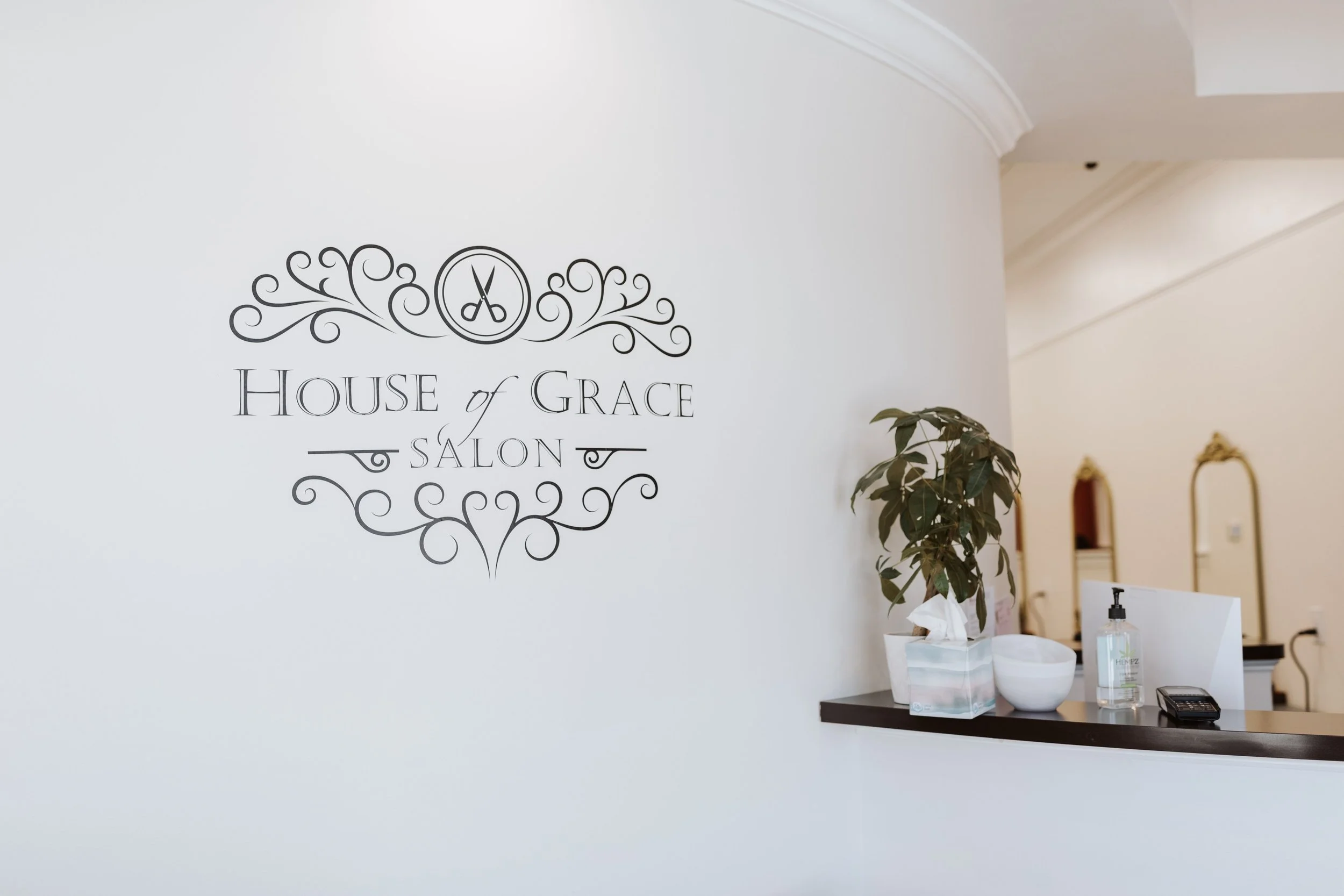 Reception area of House of Grace Salon with a wall sign featuring decorative scrollwork, scissors logo, and salon name, along with a counter holding tissues, sanitizer, and a calculator.