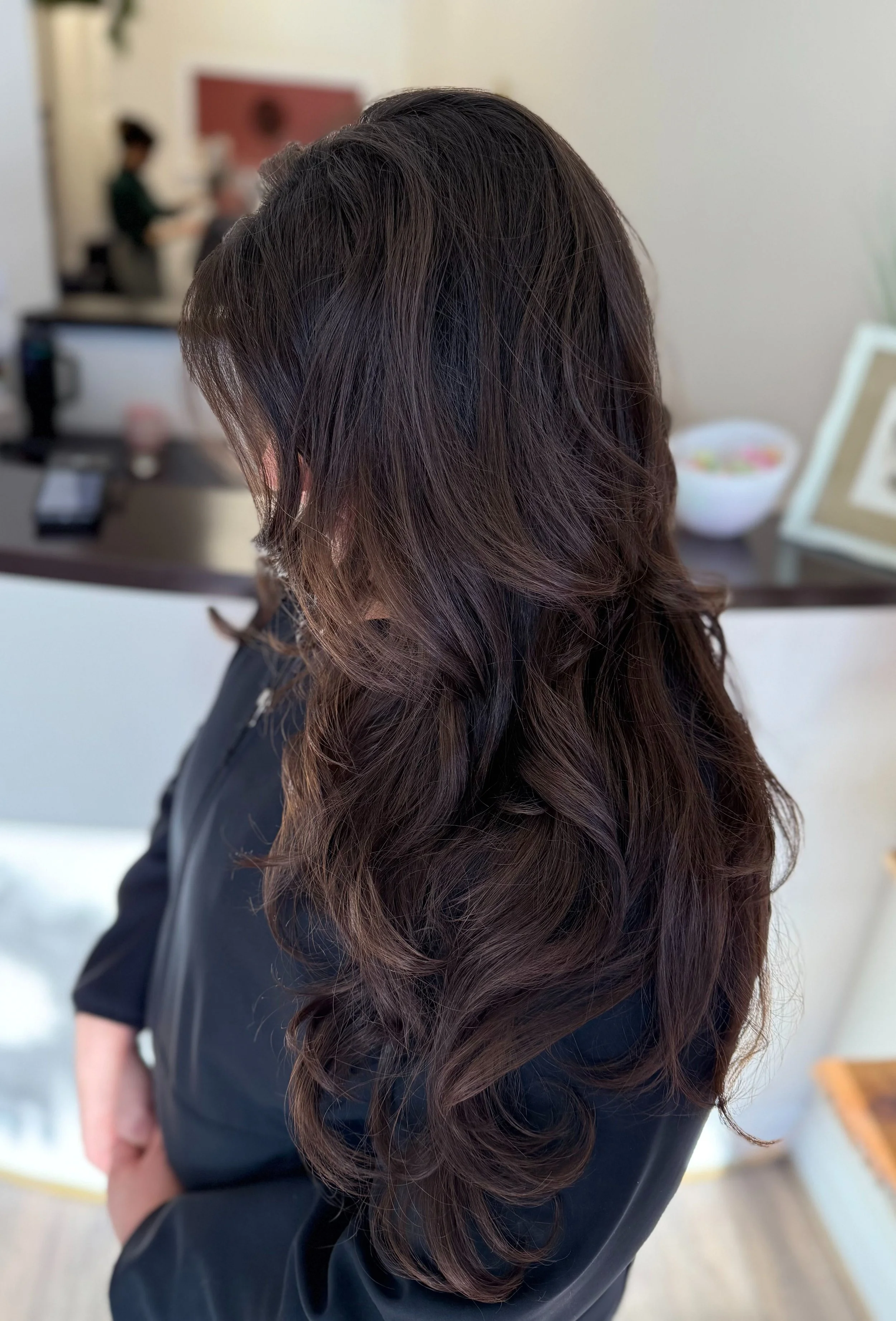Close-up of a woman with long, wavy brown hair, standing indoors with blurred background including people and furniture.