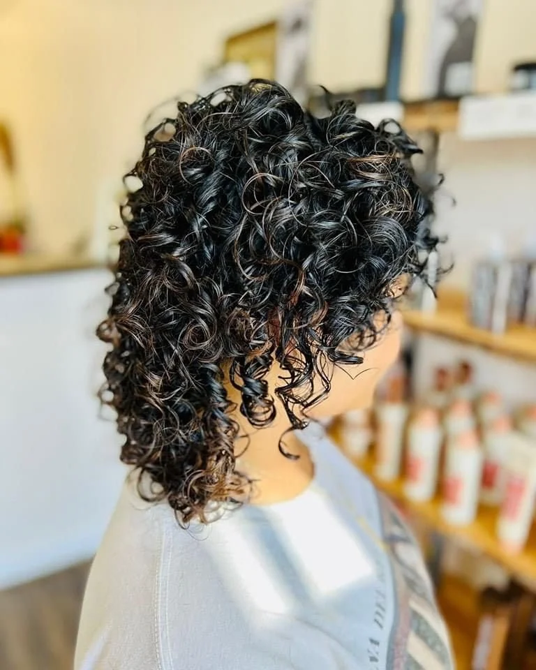 Close-up of a woman with dark, curly hair with some light highlights, in a salon setting.