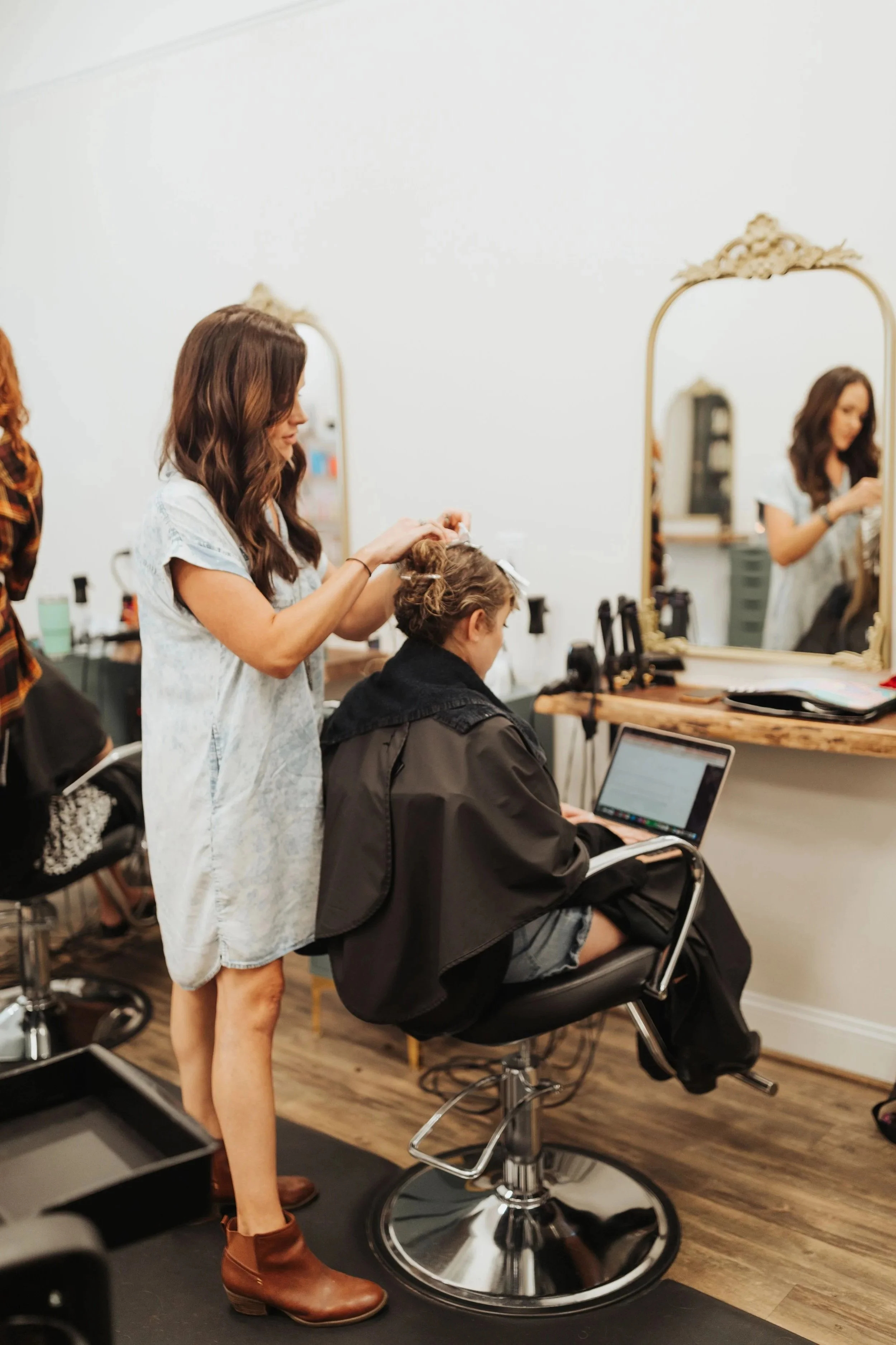 Hairdresser standing and styling a client's hair in a salon, with a large mirror and salon equipment visible in the background.