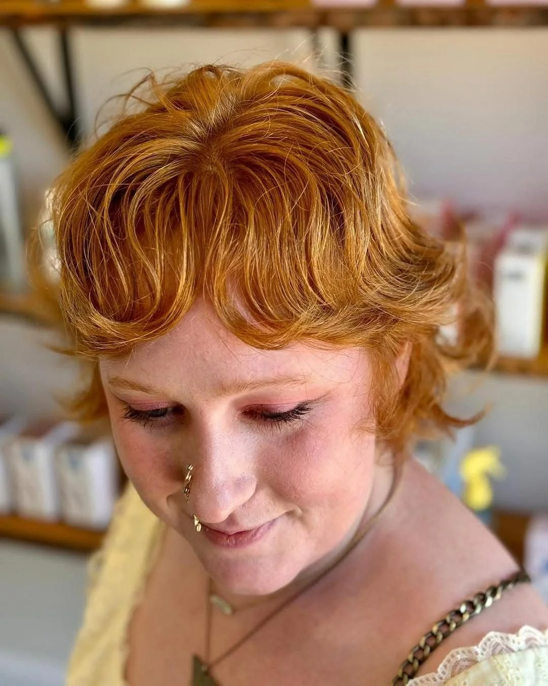 A woman with short, curly red hair looking down, with a nose piercing, wearing a yellow top with lace details and layered necklaces.