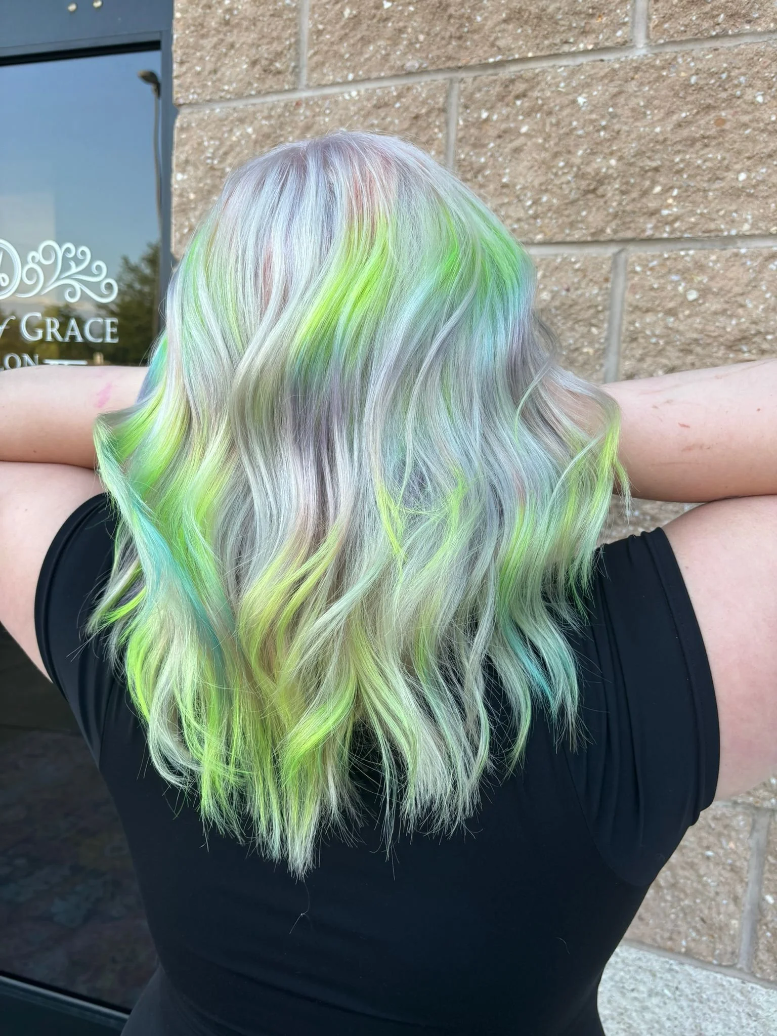 Person with long, wavy hair dyed in pastel rainbow colors, wearing a black shirt, standing against a brick wall.