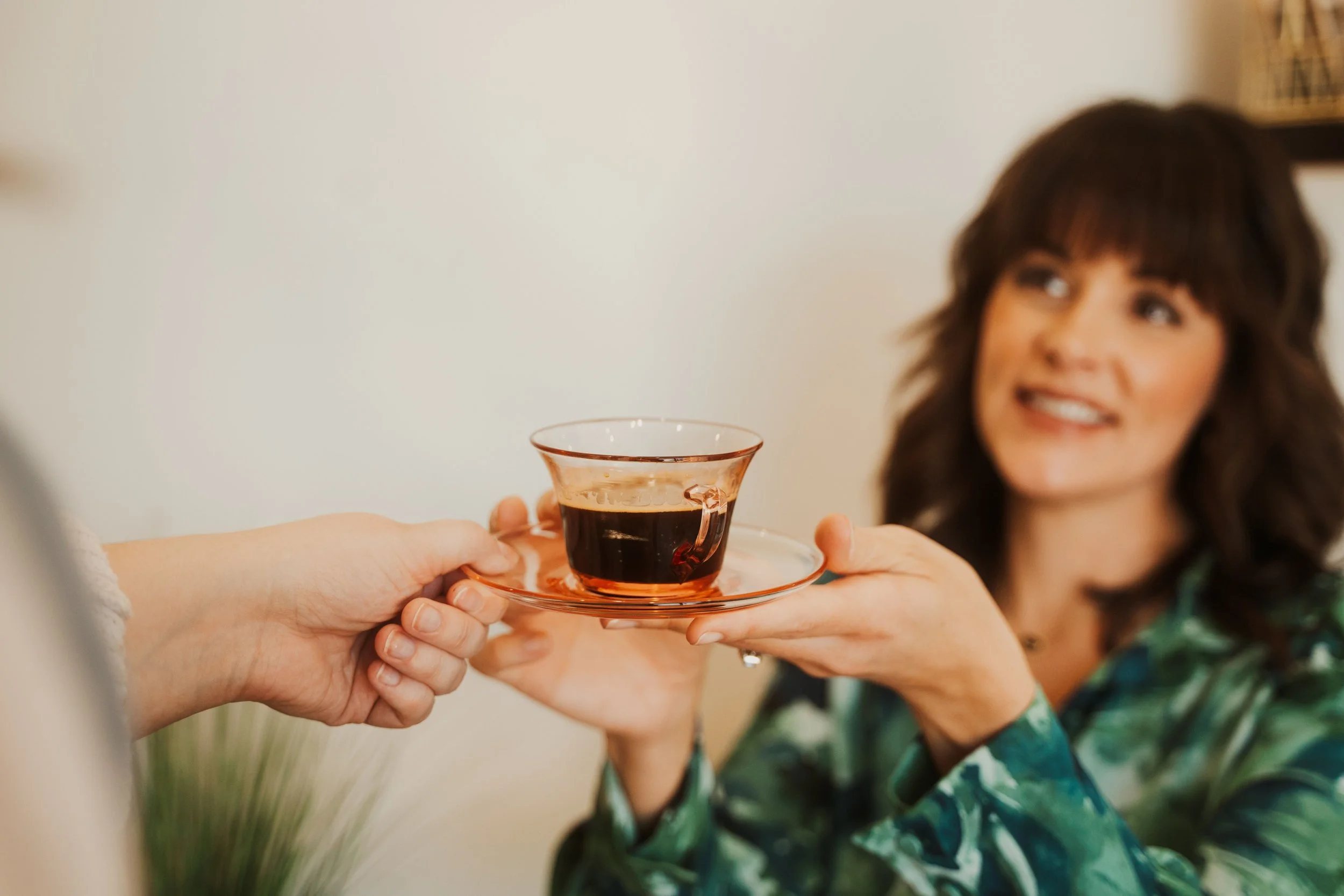 Person with wavy brown hair holding a glass cup of coffee or tea, smiling in the background.