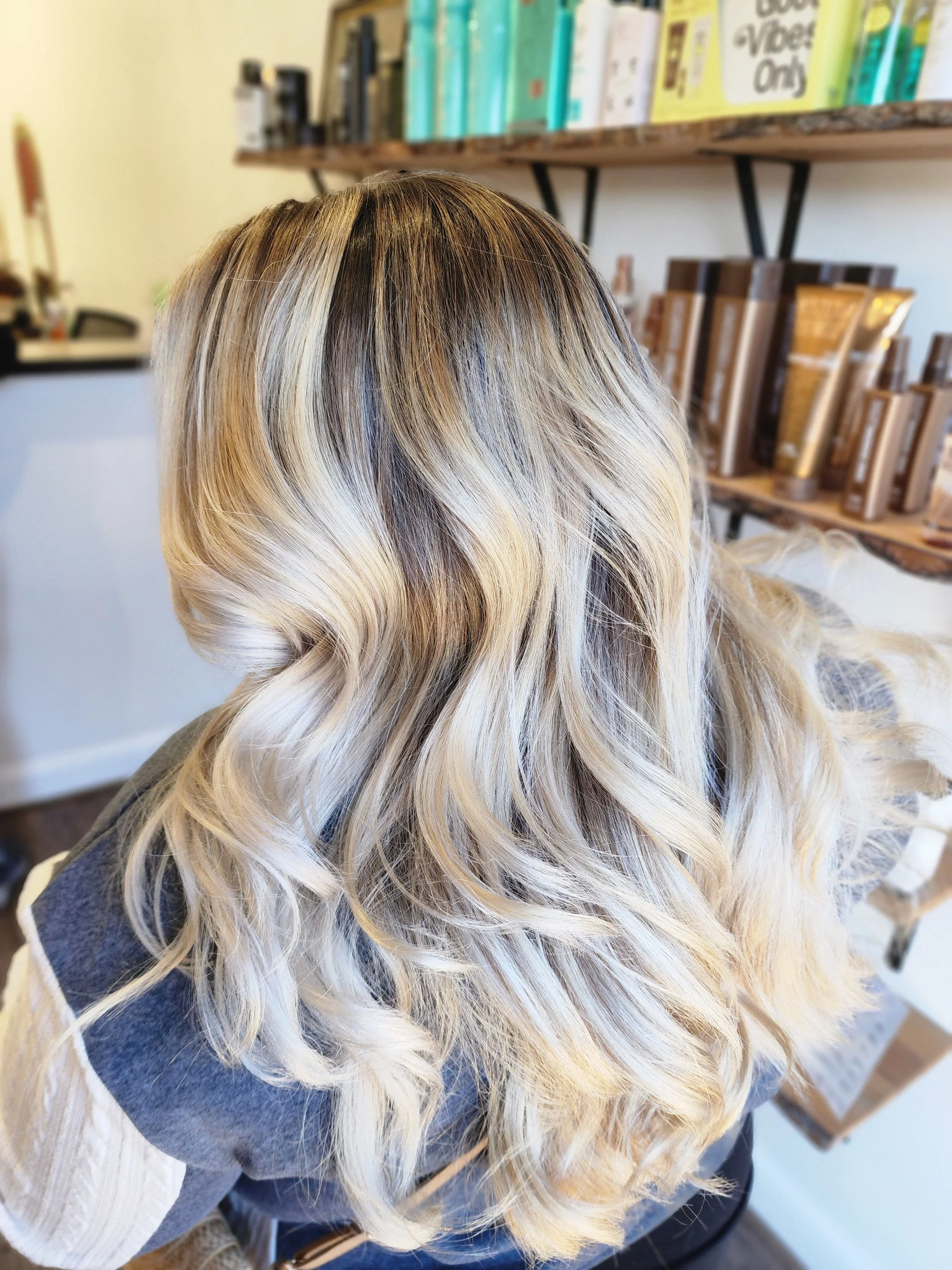 Side view of a person's head with long, wavy blonde and light brown hair, in a salon or similar setting with shelves of hair products and books in the background.