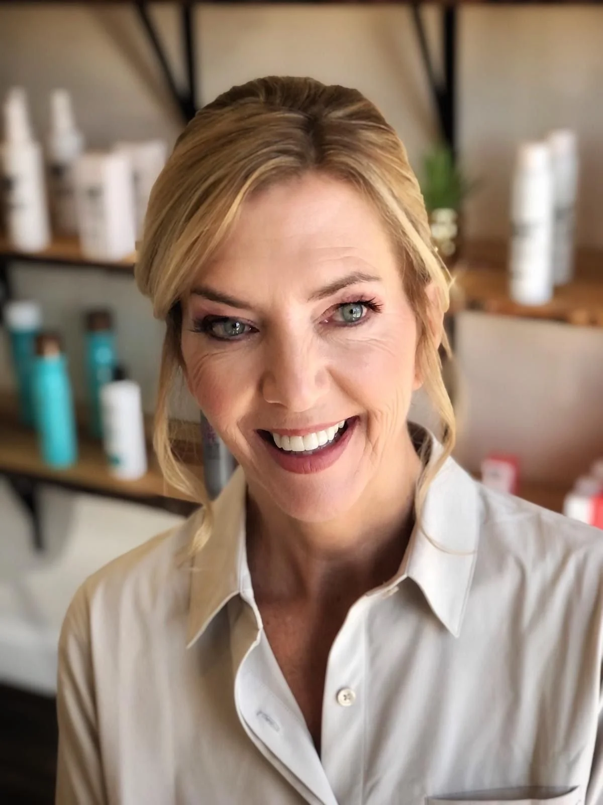 A smiling woman with blonde hair and a white shirt in a salon or spa setting with hair and skincare products in the background.