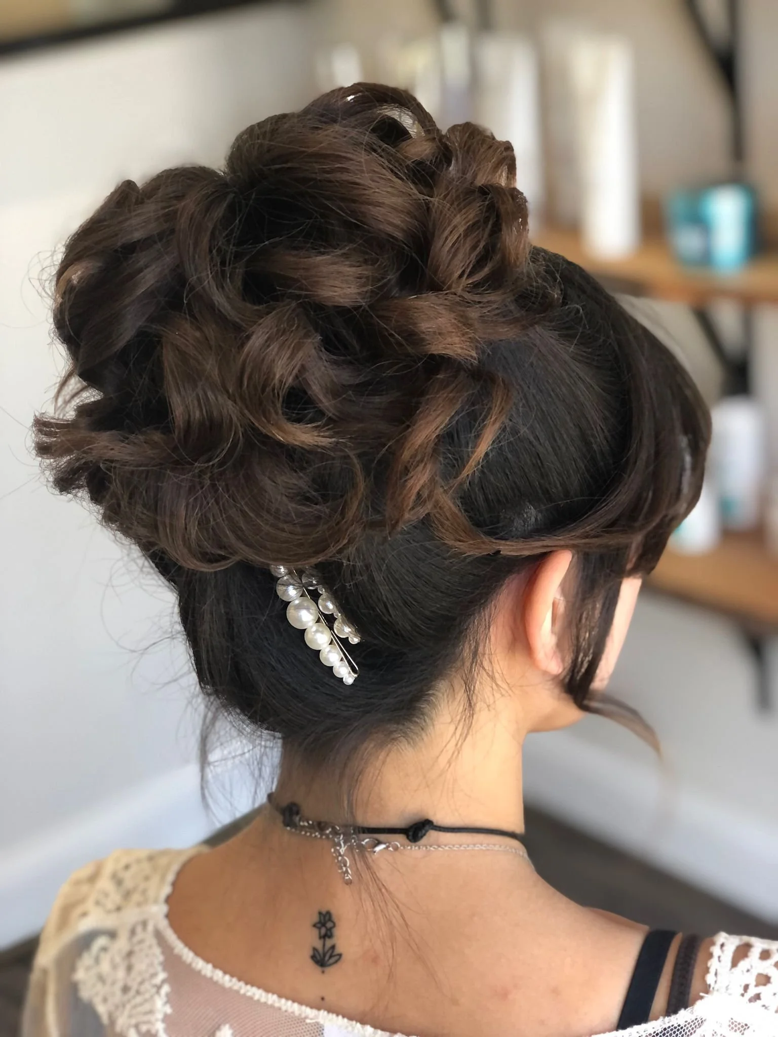 A woman's updo hairstyle with brown curls, adorned with pearl hairpins, worn with a white lace top, pearl jewelry, and a small tattoo on her shoulder.