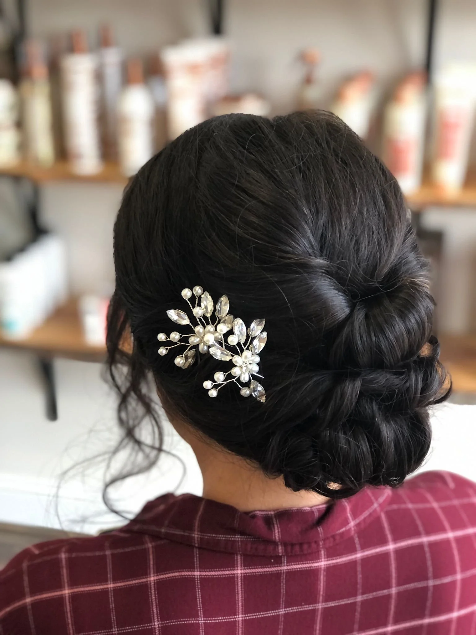 Back of a woman's head with styled dark hair decorated with a pearl and crystal floral hair accessory, wearing a maroon plaid shirt.