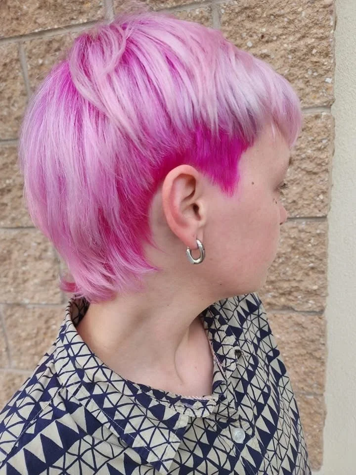 Person with short, pink and purple hair wearing a patterned shirt and a silver hoop earring, standing against a brick wall background.
