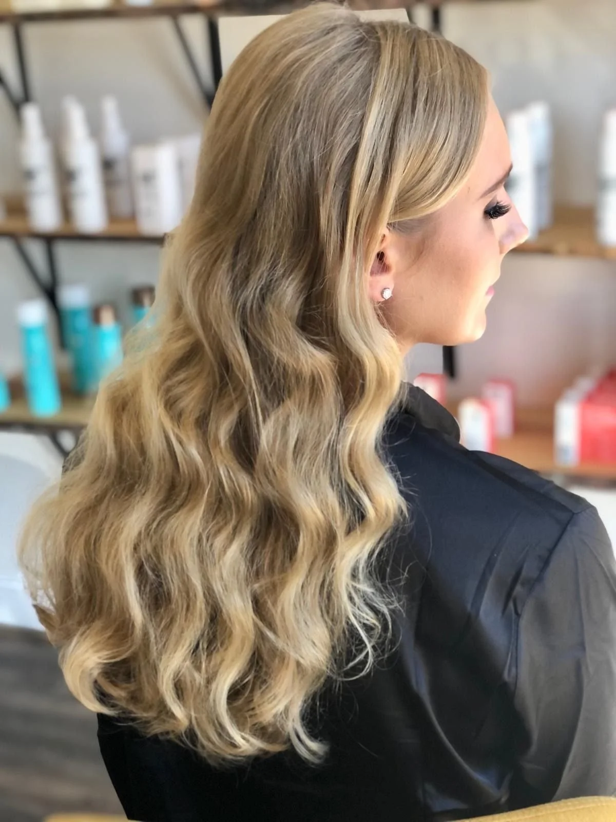 A woman with long, wavy blonde hair and small diamond earring, seen from the side, in a salon or store with hair and beauty products on shelves.
